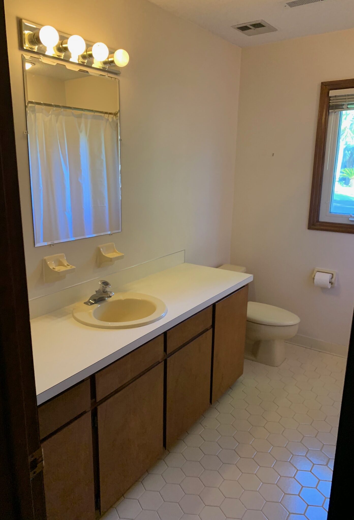 Before photo of a dated hall bathroom featuring yellowed tiles, warm wood cabinets, and an outdated vanity light. The space has a worn, vintage look reminiscent of an old motel, in need of a modern renovation.