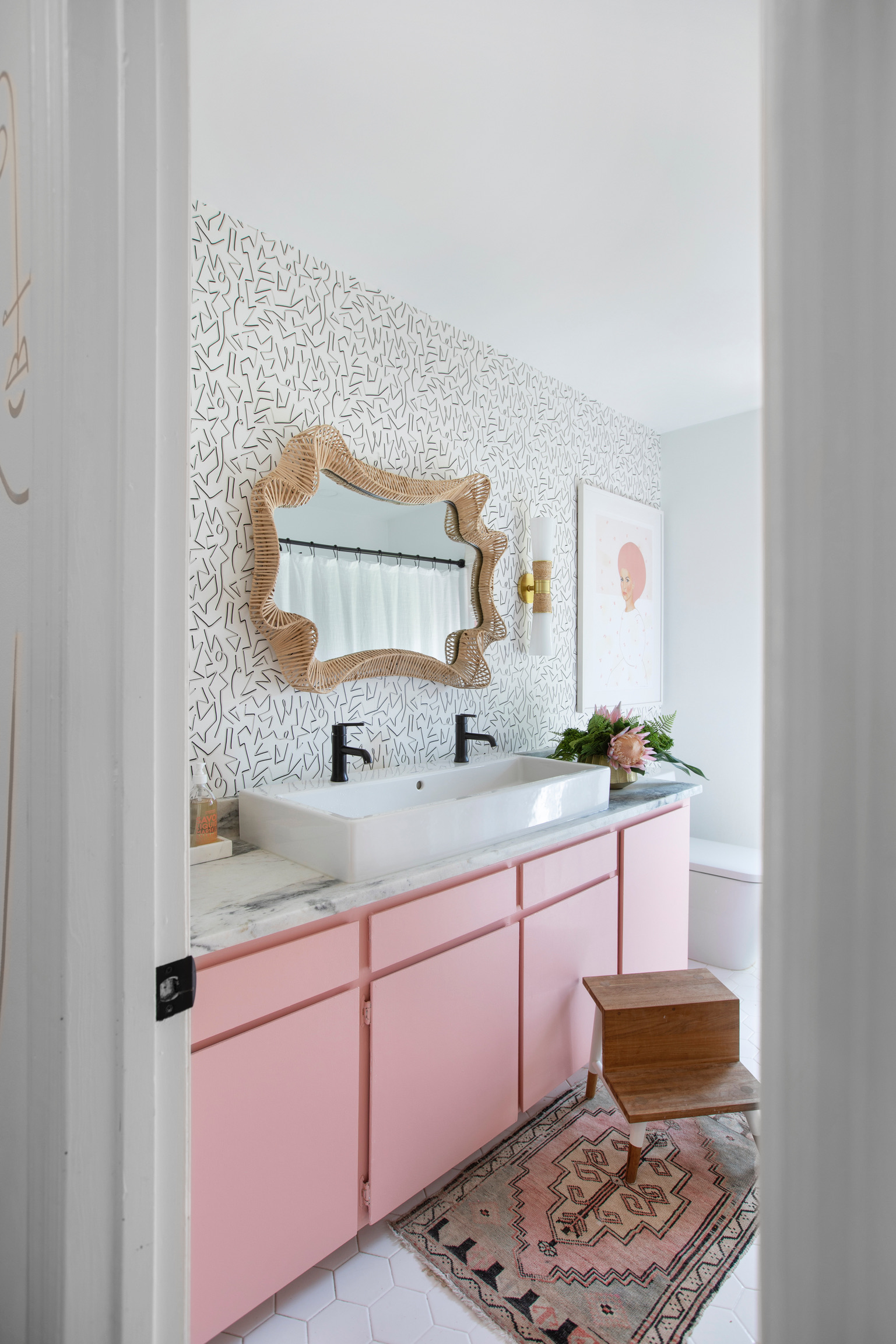 After photo of a glamorous and girly hall bathroom renovation featuring original cabinets painted bubblegum pink, a funky patterned wallpaper accent, and a unique woven mirror. A modern white trough sink replaces the old vanity, complemented by Megan Molten’s exclusive Mitzi sconces. The original tiles remain but have been refreshed with crisp white paint, creating a fun, stylish, and perfectly modern space for little girls.