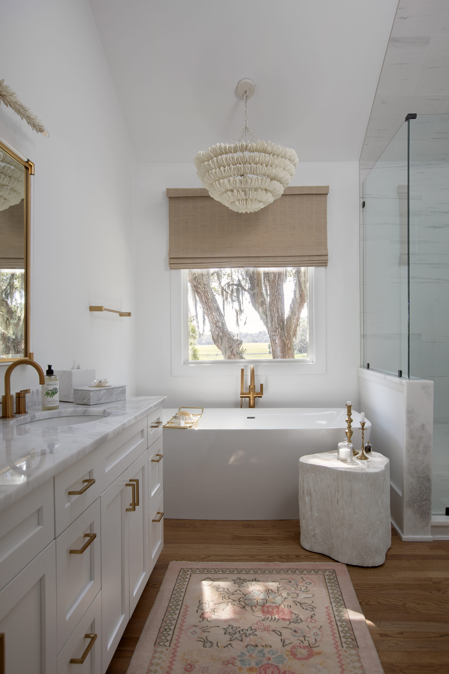 Coastal luxury bathroom designed by Megan Molten featuring a freestanding soaking tub, marble vanity, brass fixtures, custom cabinetry, woven chandelier, and natural light overlooking the Charleston Lowcountry.