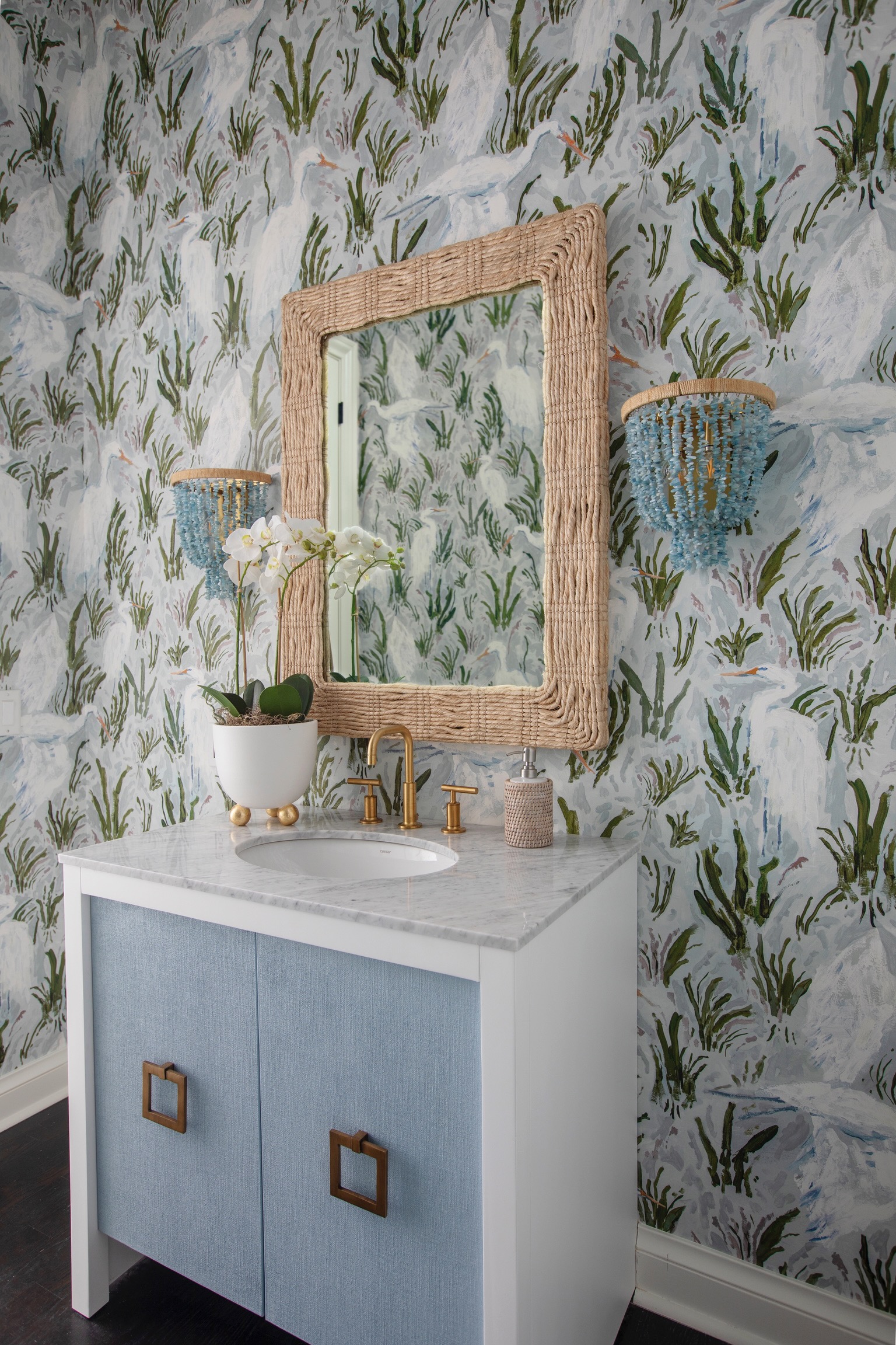 Coastal powder room with grasscloth mirror frame, blue vanity with brass hardware, marble countertop, and patterned wallpaper in a Charleston designer home.