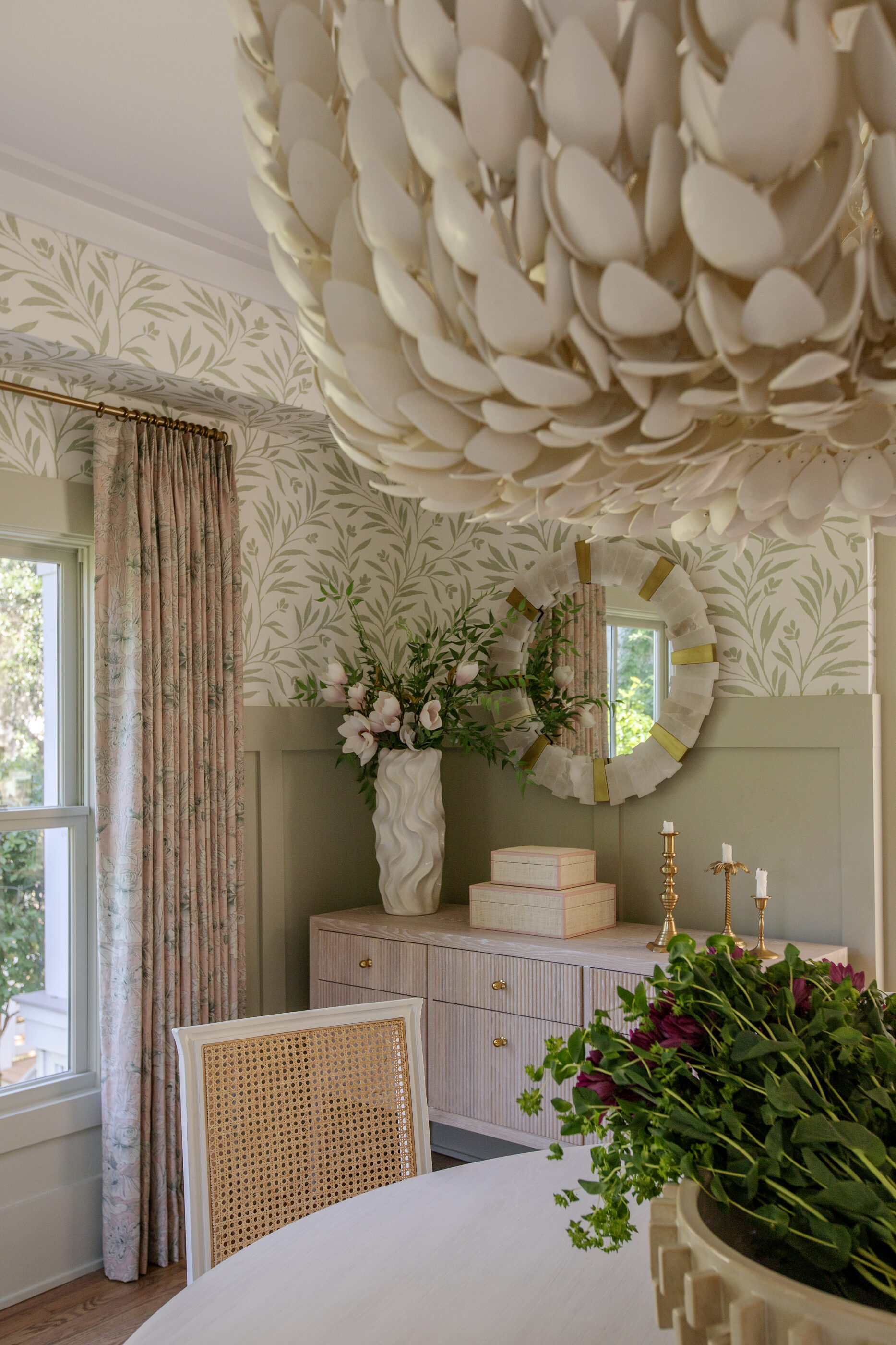 Coastal dining room with soft green wallpaper, blush drapery panels, cane dining chair, sculptural chandelier, and custom built-in sideboard styled in a modern Southern interior design.