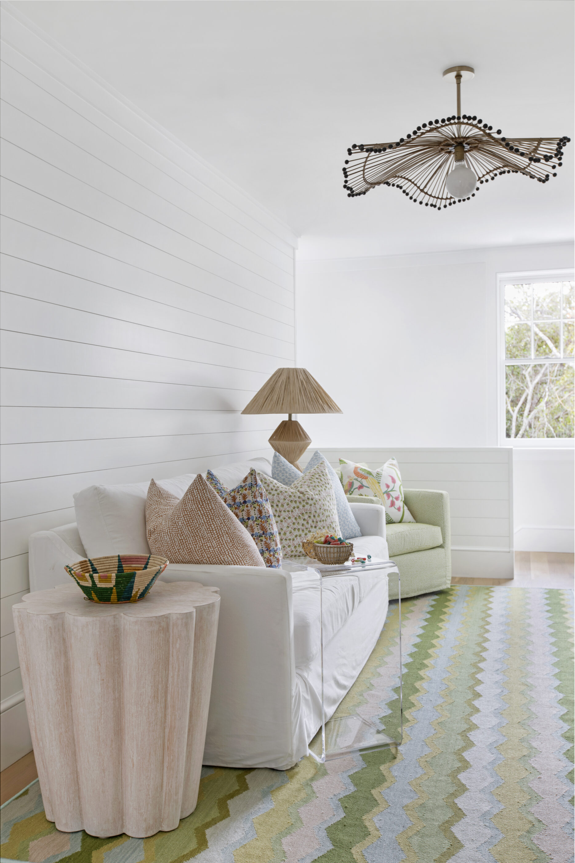 Bright coastal sitting room with white shiplap walls, neutral sofa, colorful patterned pillows, woven table lamp, pastel zigzag rug, and modern ceiling light fixture.