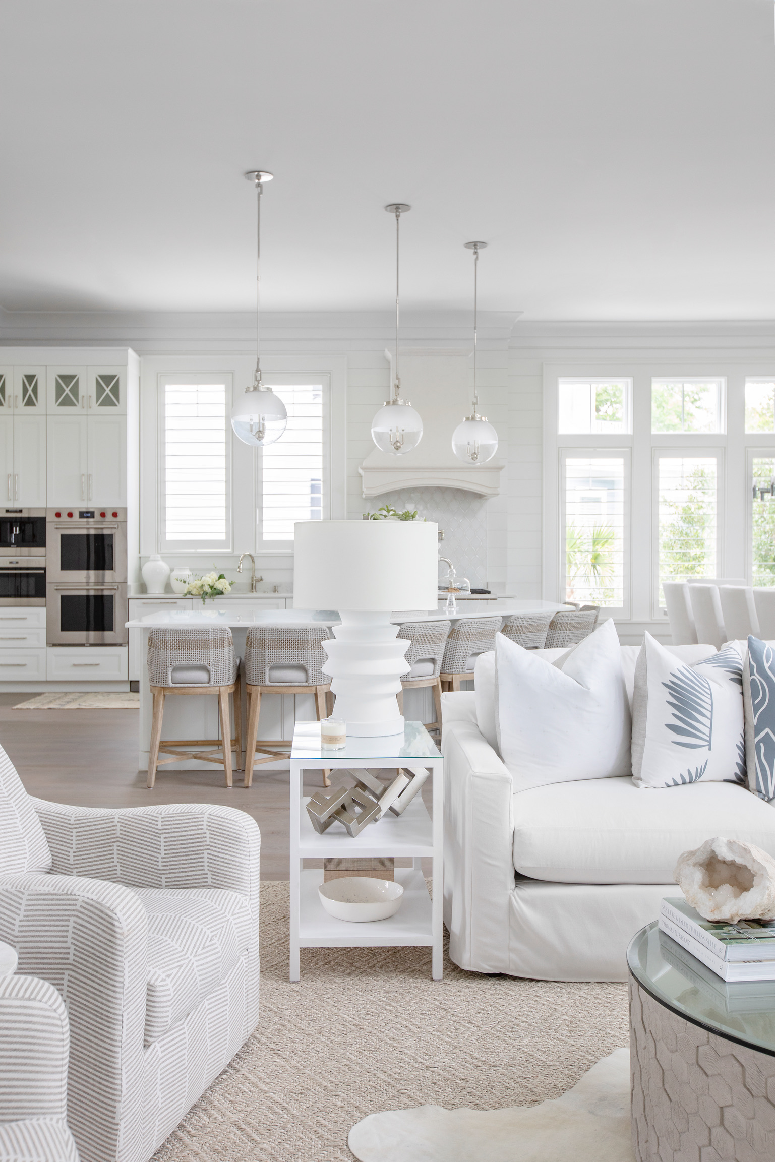 Bright, airy coastal living room and kitchen with an all-white palette, featuring a slipcovered sofa, textured armchairs, and a woven rug. A glass and metal side table with a sculptural white lamp sits between seating, while the open kitchen in the background showcases white cabinetry, barstools, and three pendant lights over the island. Large windows and soft natural textures create a clean, relaxed feel.