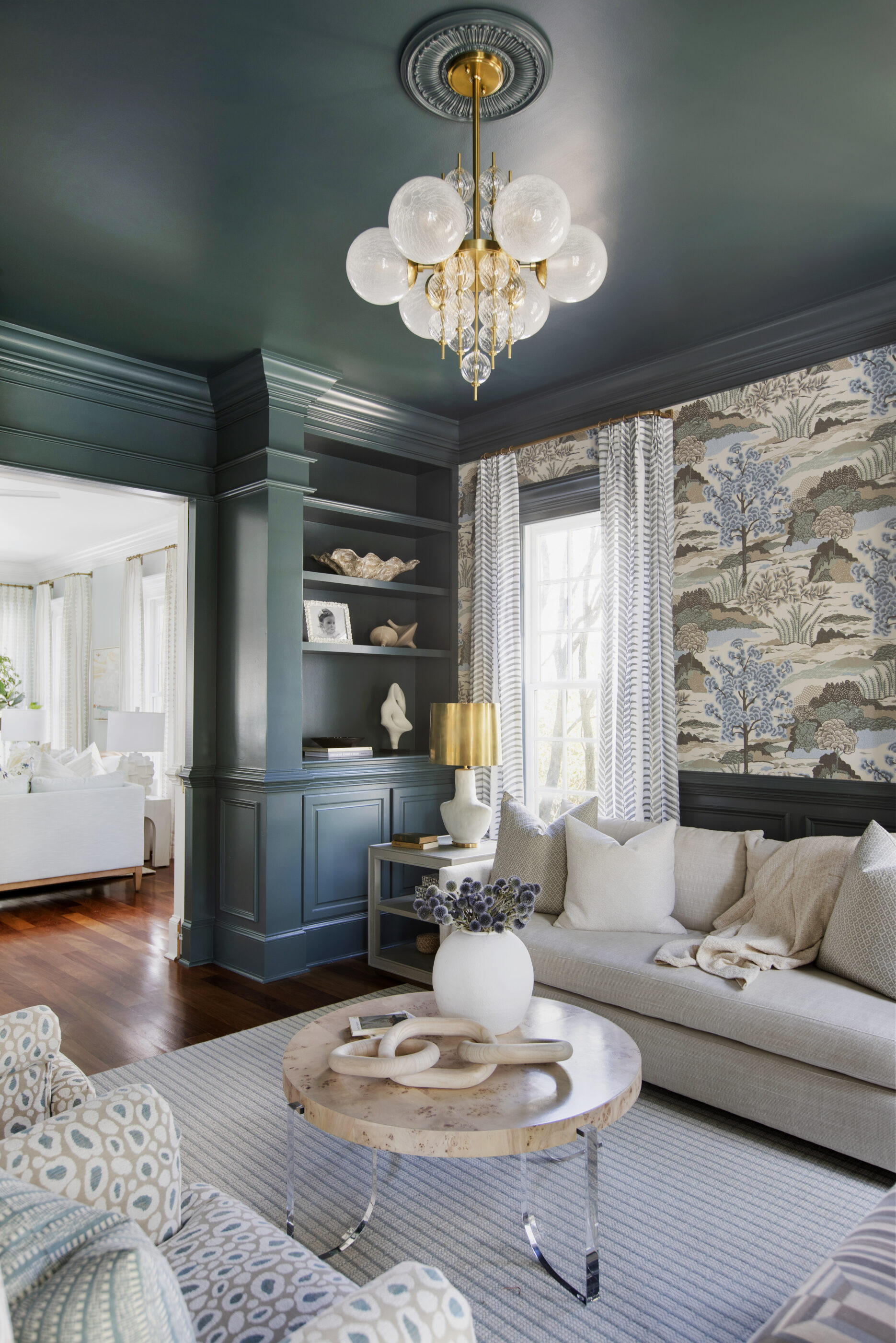 Coastal-inspired living room with deep green built-in shelving and ceiling, patterned wallpaper accent wall, and a modern brass chandelier with glass globes. A neutral sofa with layered throw pillows sits beside a light wood and acrylic coffee table, styled with a ceramic vase and decor pieces. Natural light filters through striped curtains, highlighting warm wood floors and a soft textured rug.