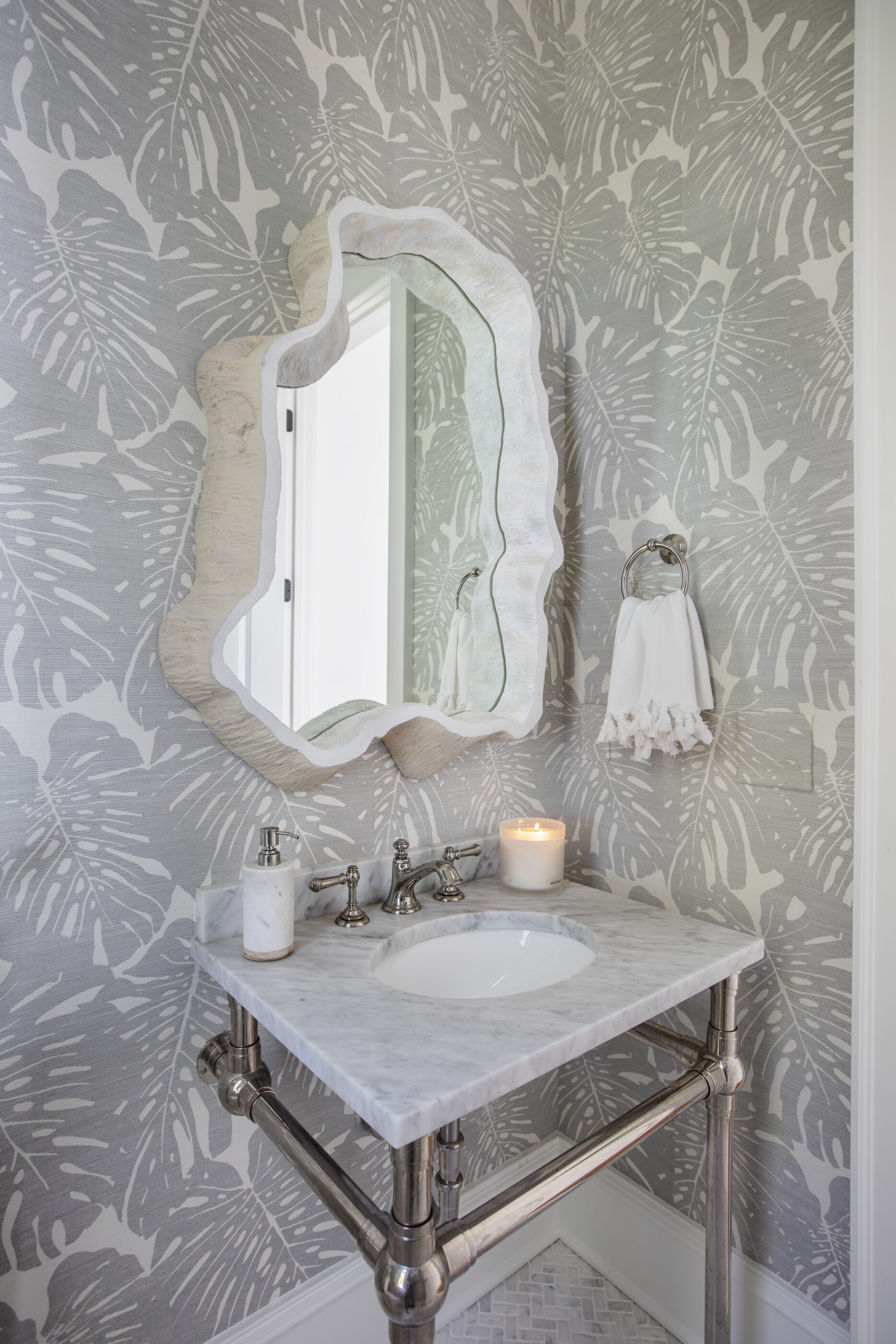 Coastal powder bathroom with white Carrara marble sink, chrome fixtures, sculptural mirror, and gray palm leaf wallpaper