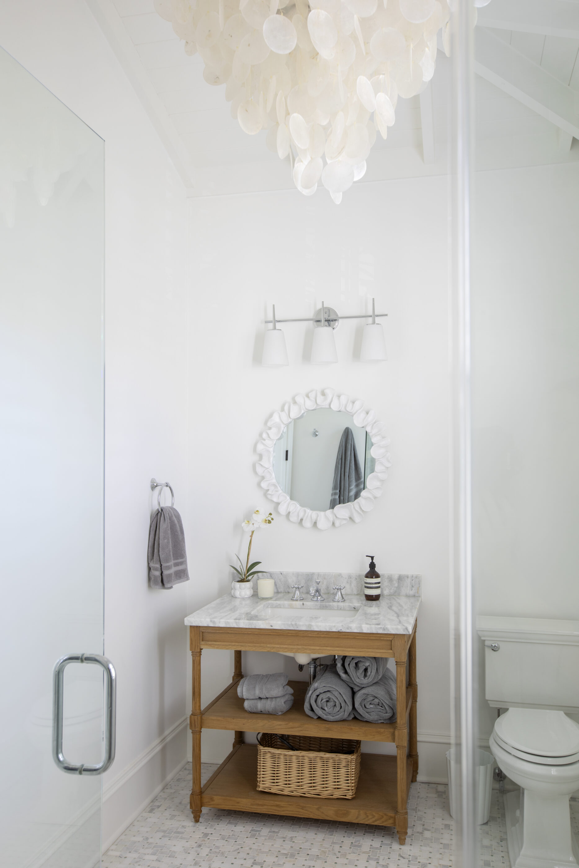 Bright coastal bathroom with Carrara marble countertop, wood vanity, glass shower, chrome fixtures, and capiz shell chandelier