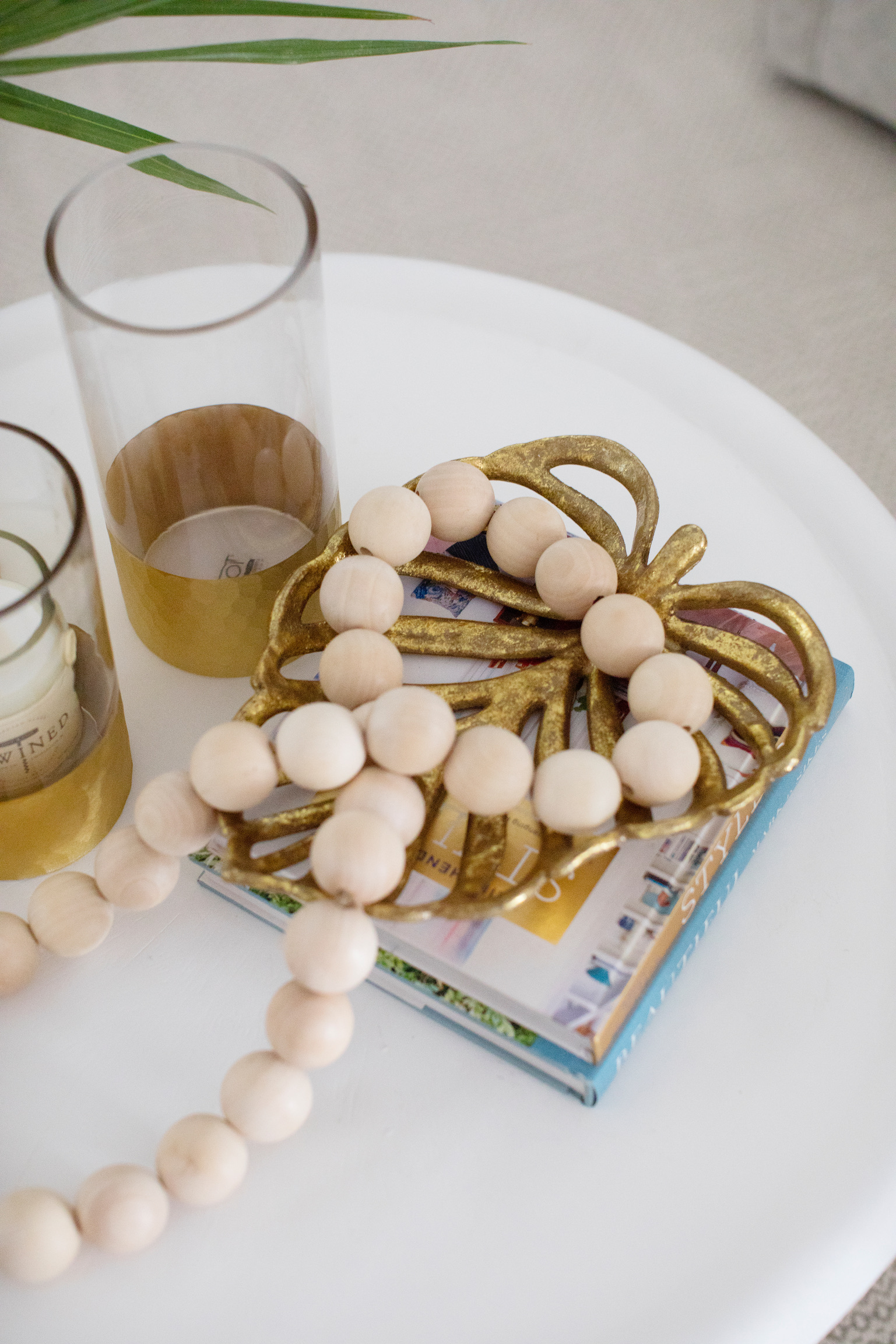 Close-up of coffee table decor with wooden beads, a gold decorative tray, glass hurricanes, and stacked design books