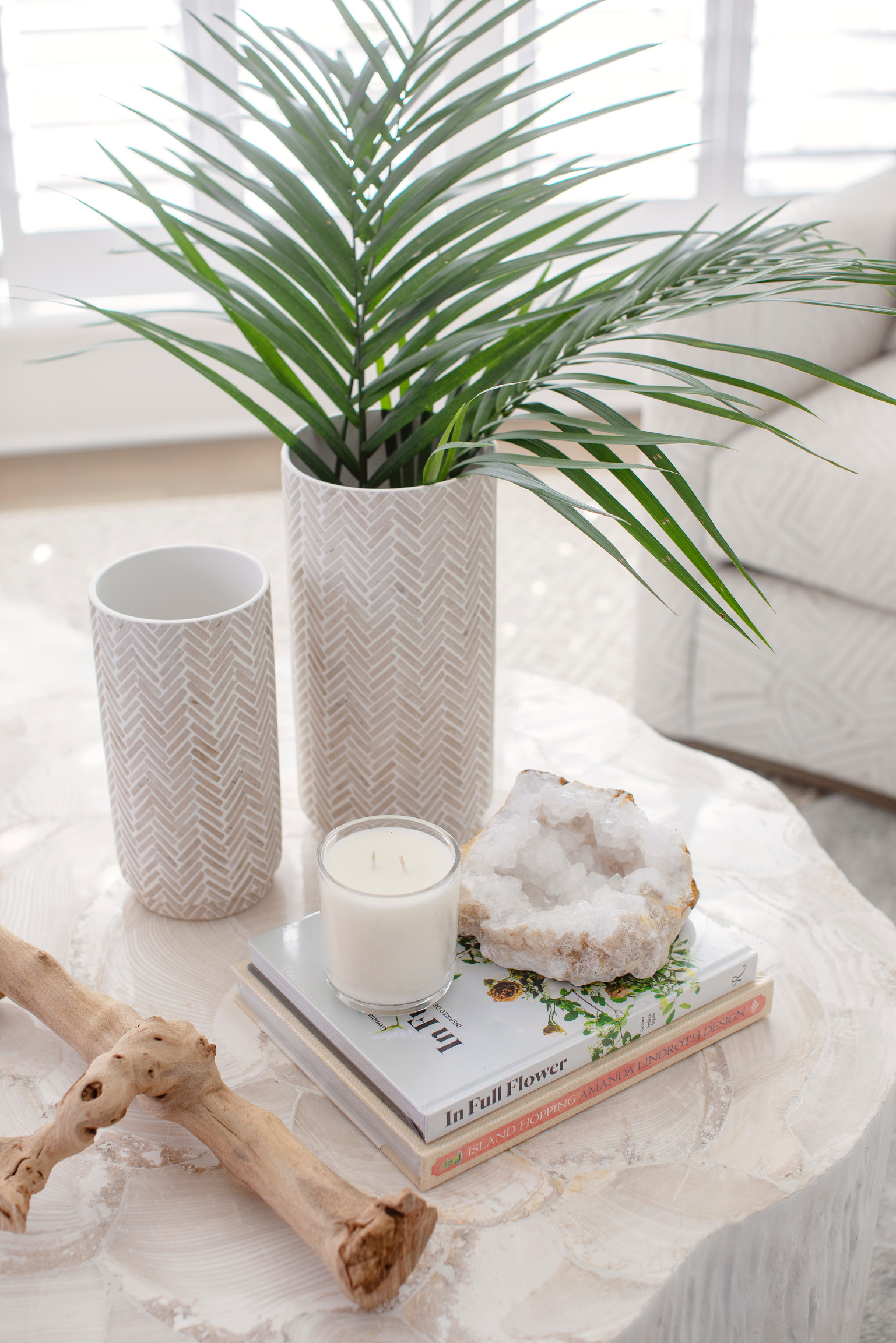 Modern coastal coffee table styled with palm leaves, ceramic vases, a candle, stacked books, driftwood, and a crystal accent