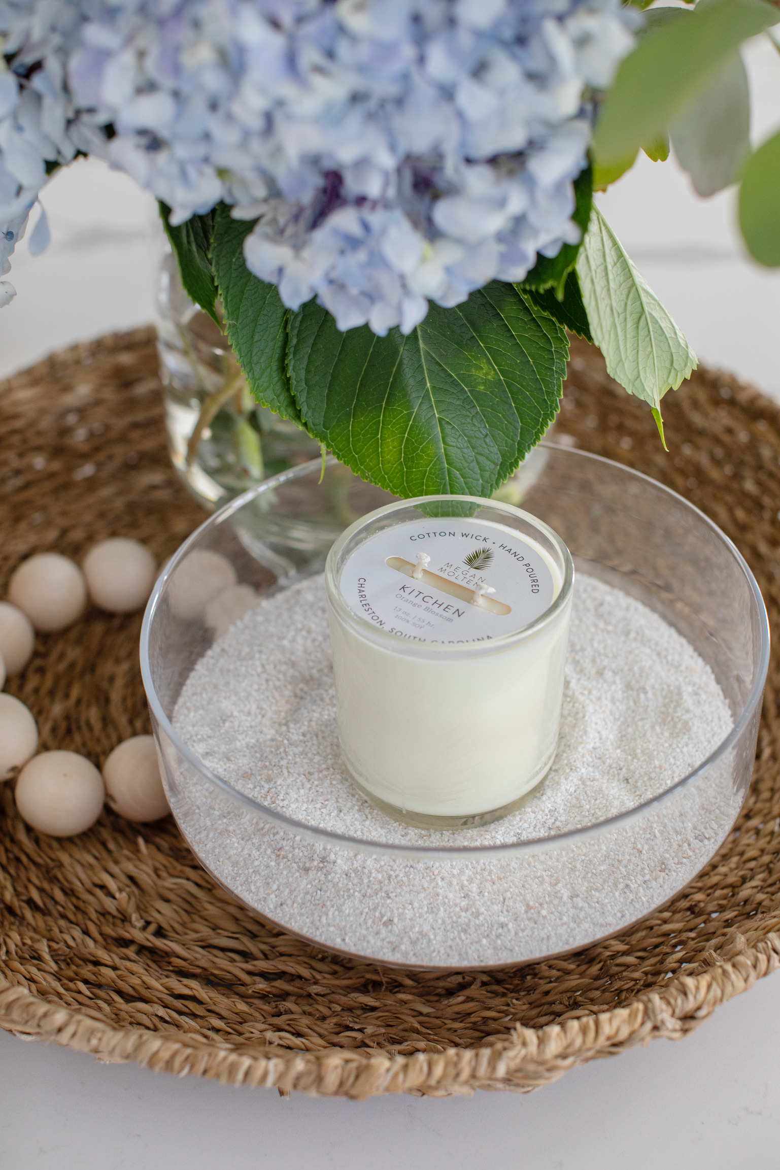 Woven tray styled with a glass candle, white sand, wooden beads, and blue hydrangeas for a coastal coffee table look