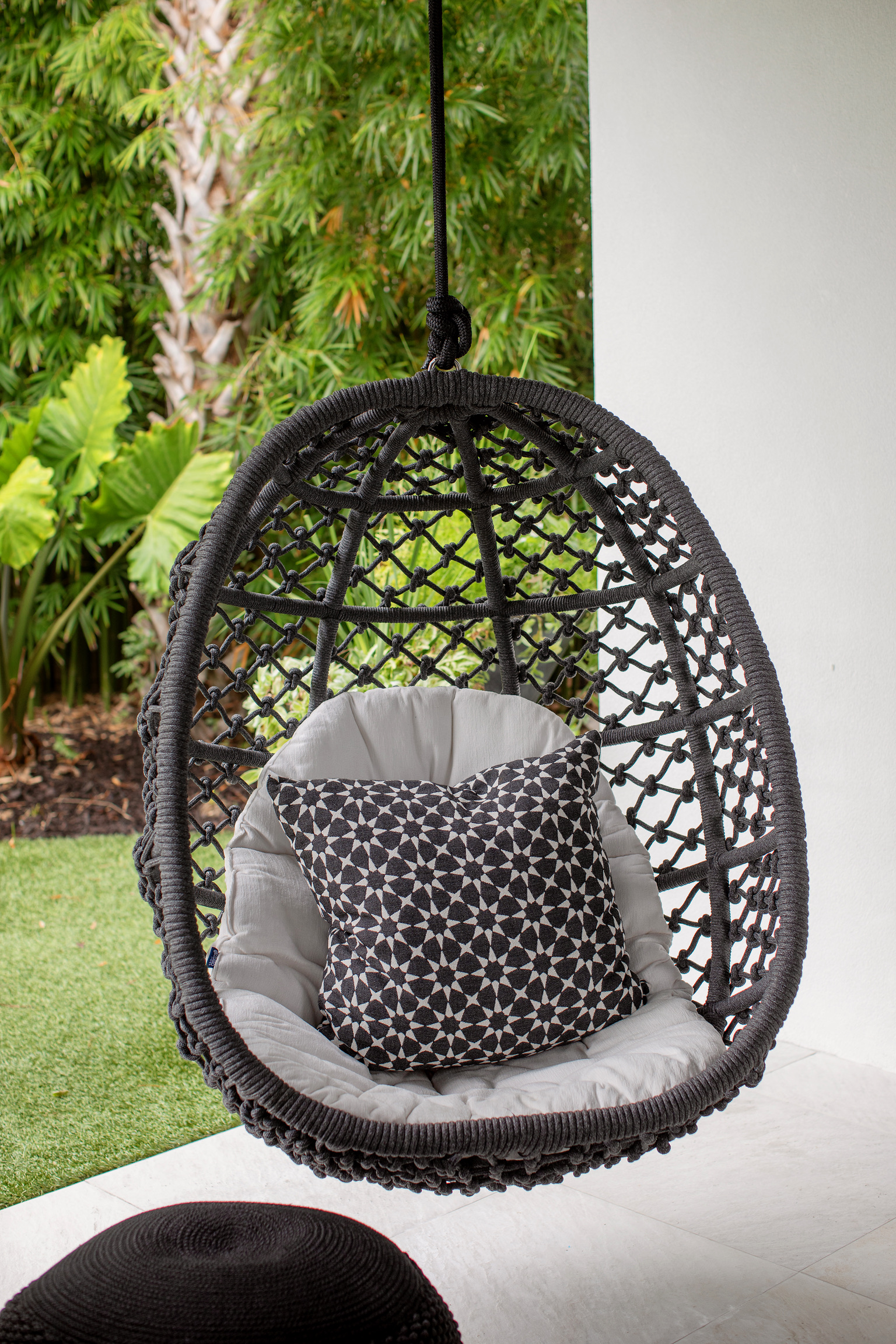Black hanging egg chair with patterned designer pillow in a luxury coastal cabana surrounded by tropical greenery.