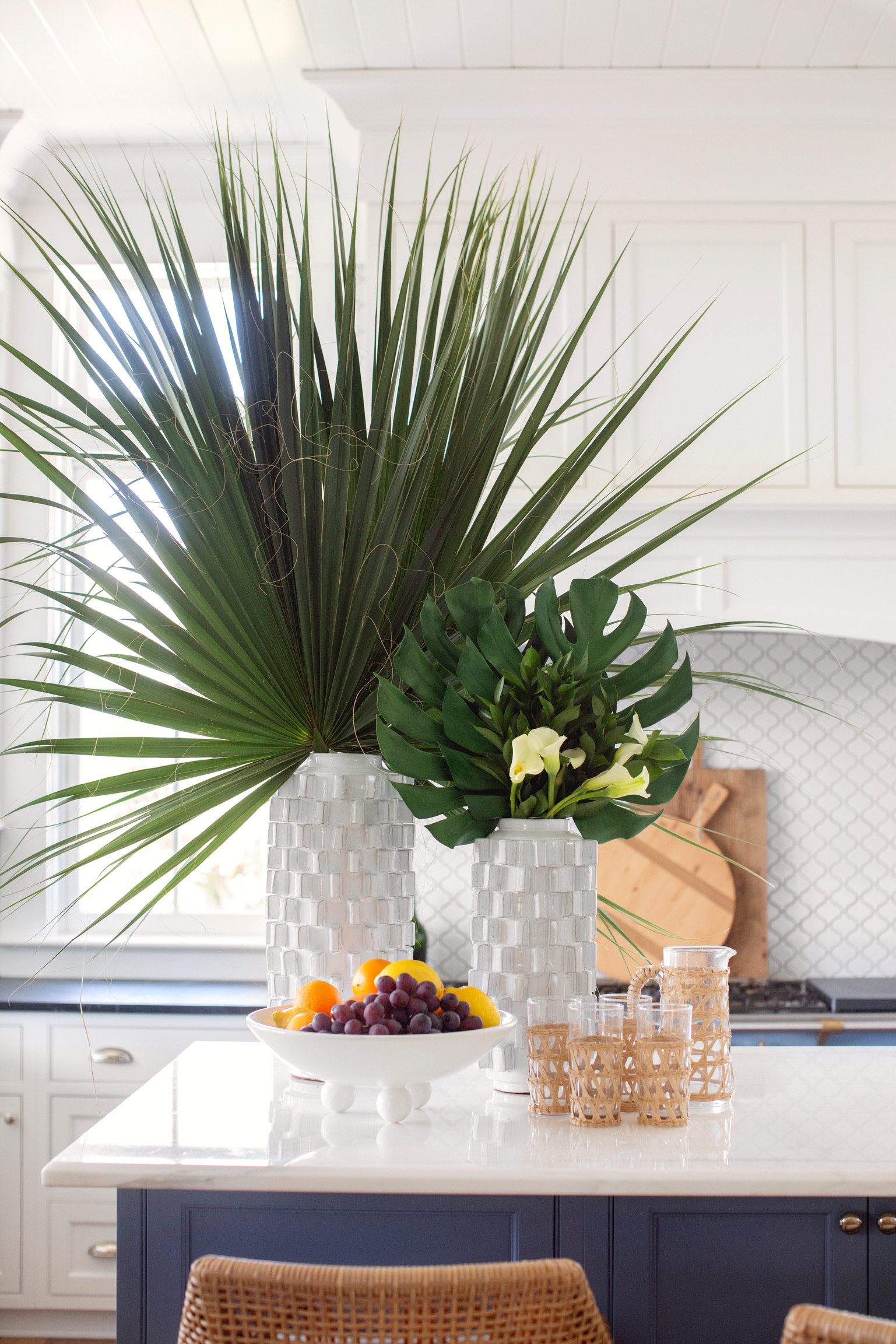 Kitchen island with large tropical greenery, white textured vases, fruit bowl, and woven glassware in a bright coastal kitchen.