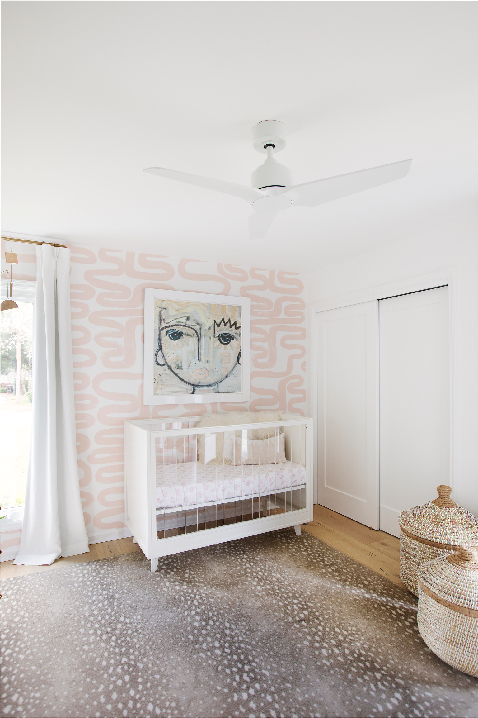 Pink nursery with white crib, abstract wall art, patterned wallpaper, white curtains, and woven baskets.
