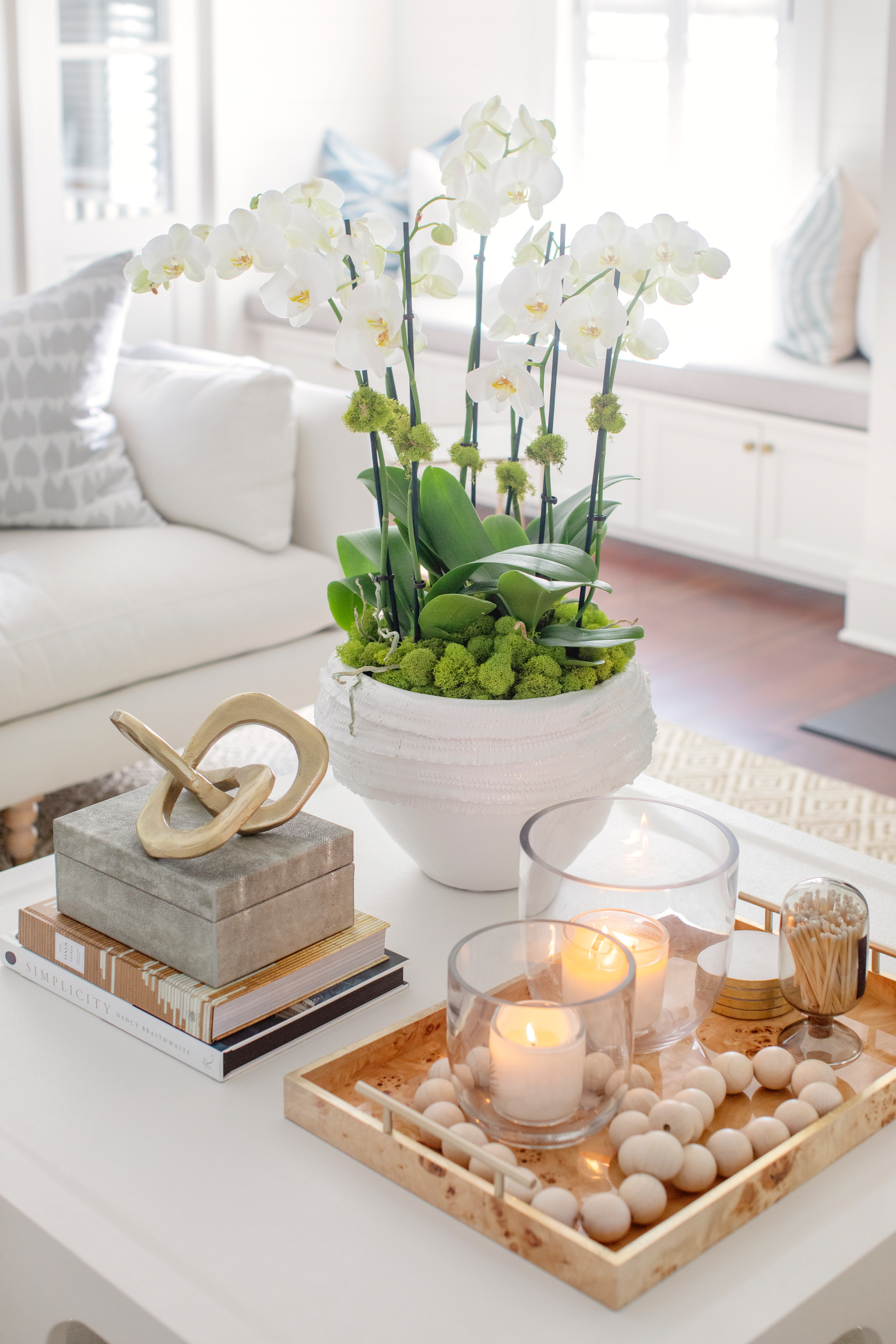 Styled coffee table with white orchids, candles, decorative beads, stacked books, and neutral coastal home accessories.