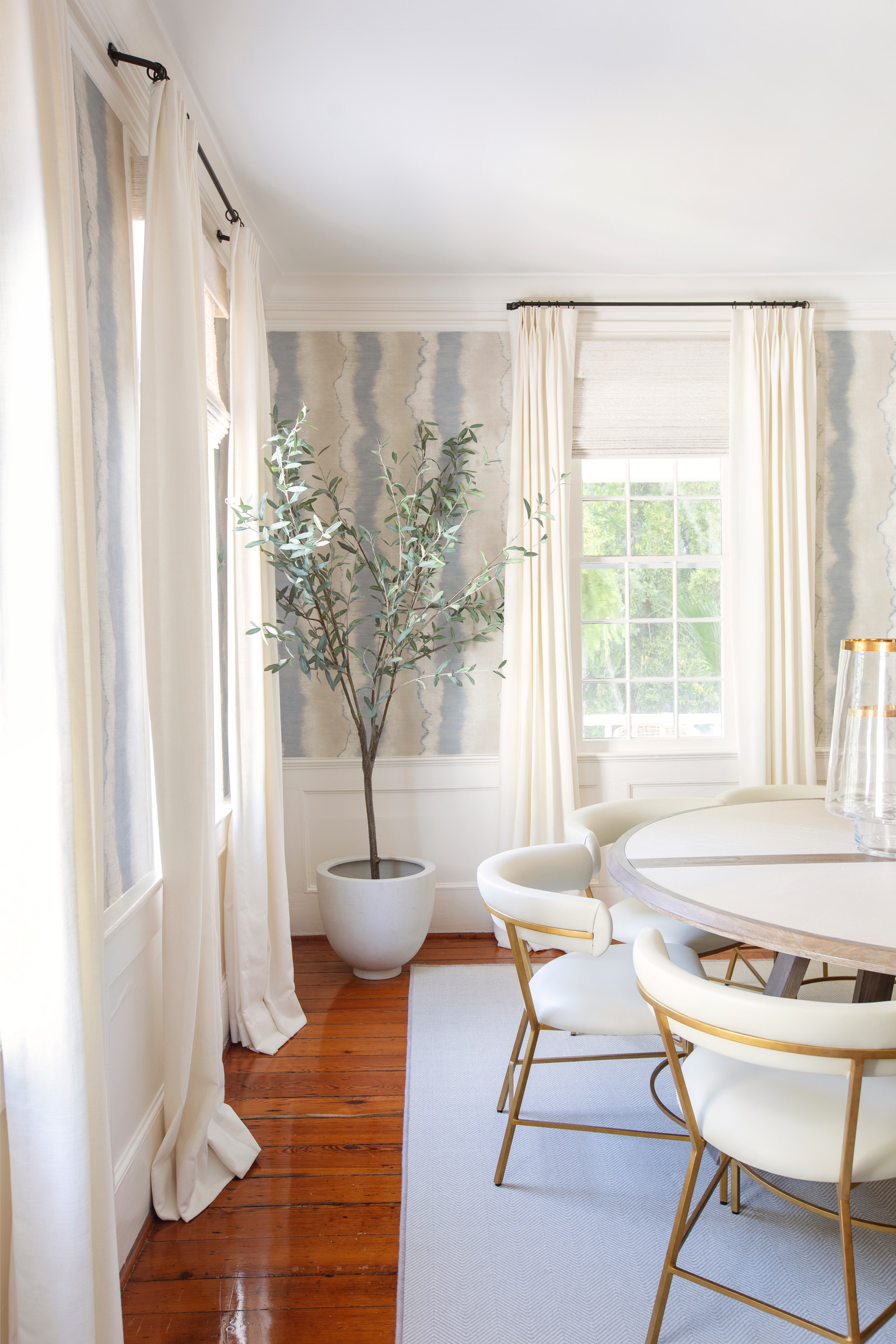 Bright dining room with patterned wallpaper, white curtains, round dining table, gold chairs, and a potted olive tree.