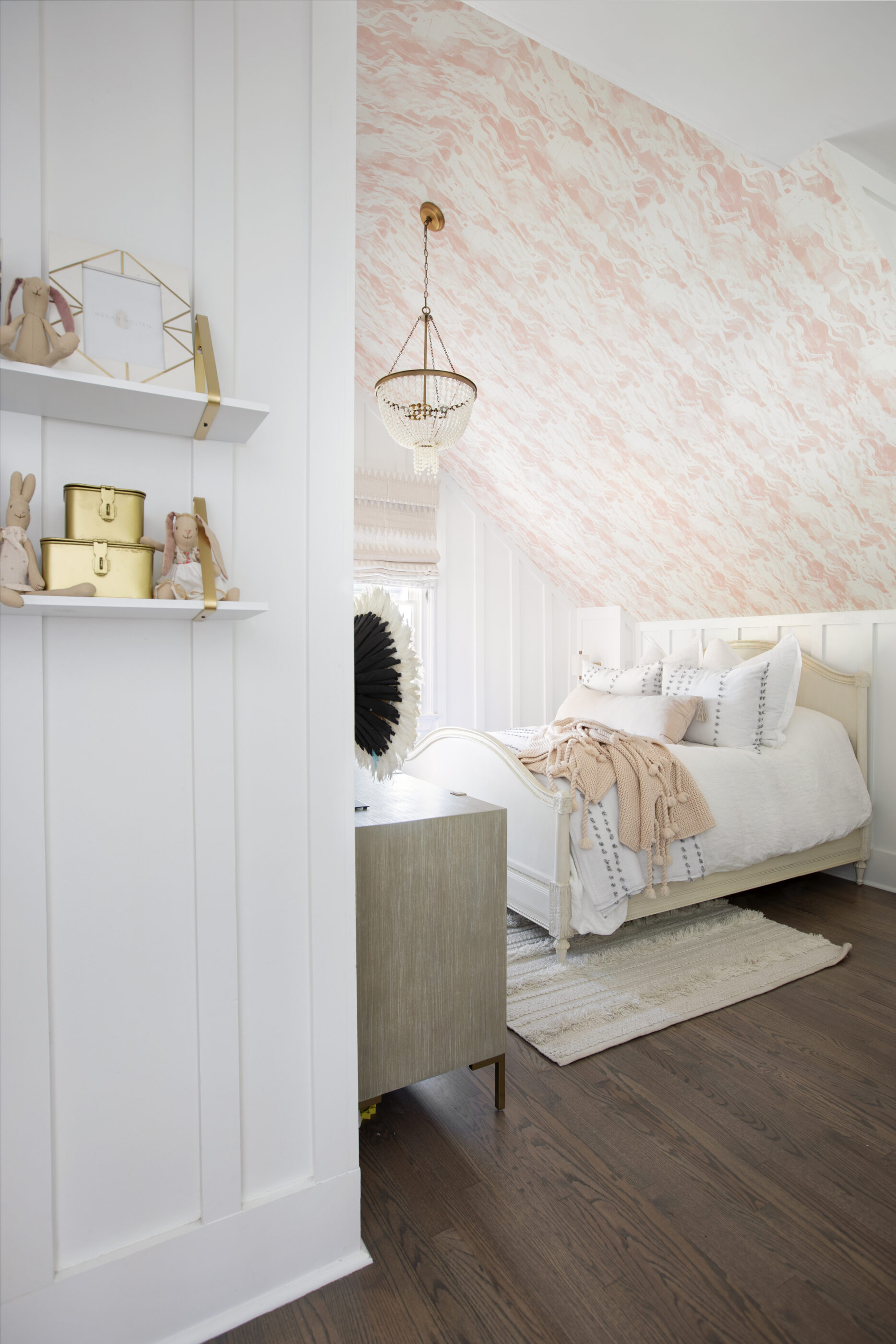 Girls bedroom with pink patterned wallpaper, white bedding, blush throw blanket, chandelier, and styled shelves.