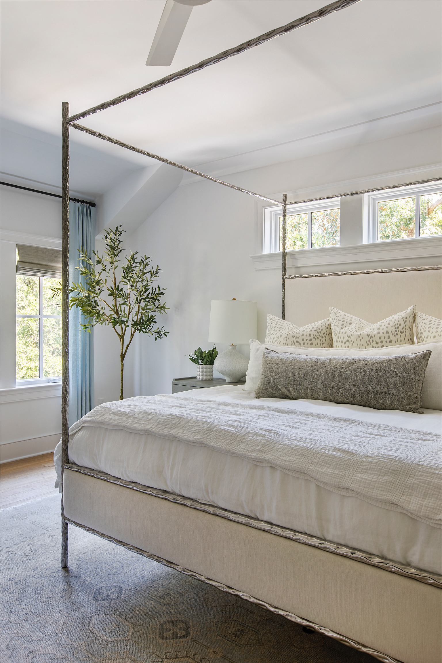 Neutral bedroom with metal canopy bed, layered bedding, patterned pillows, blue curtains, and natural light.