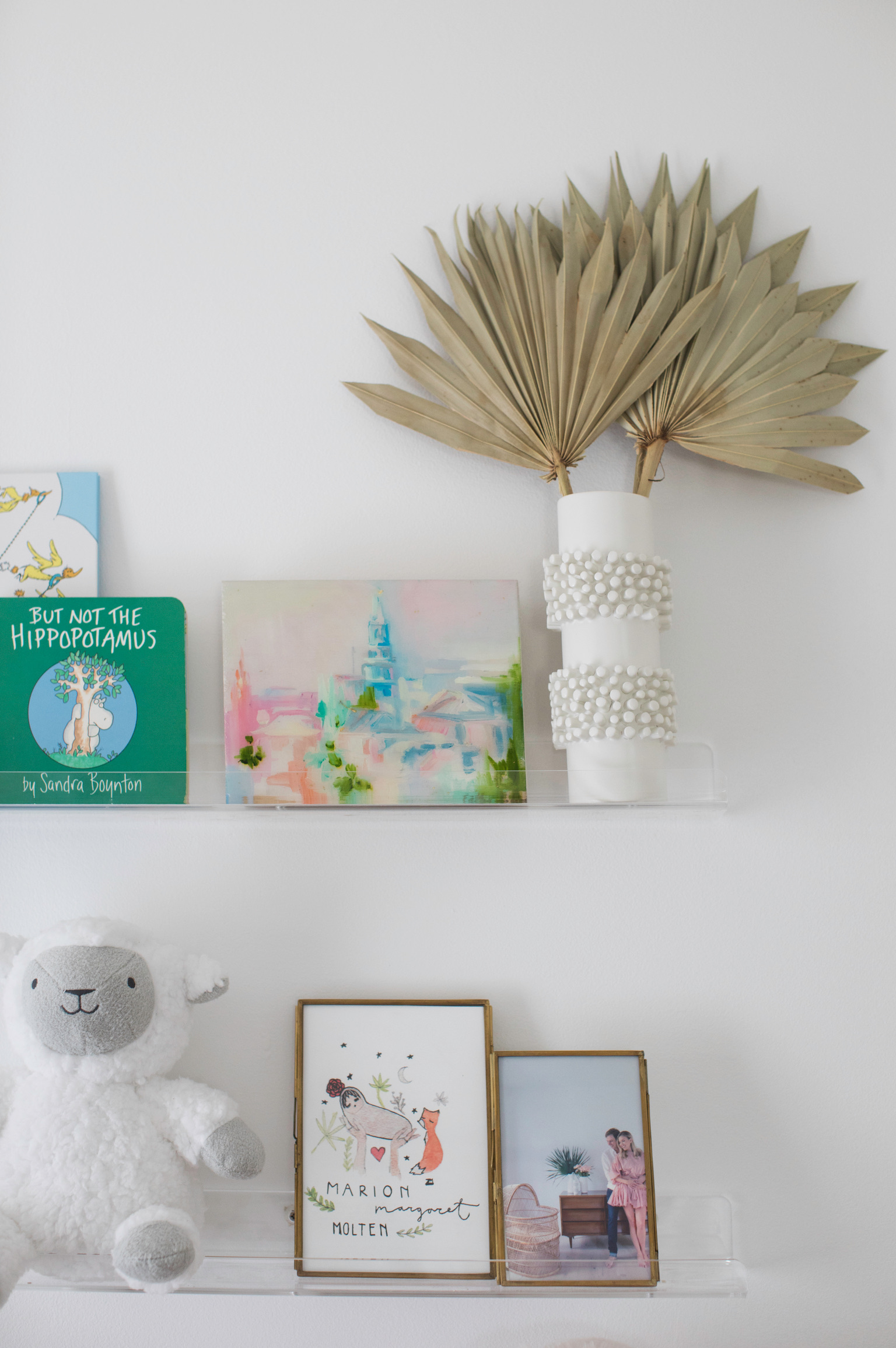 Kids room shelf styling with children’s books, framed nursery art, family photo, white textured vase, dried palm leaves, and plush lamb decor.