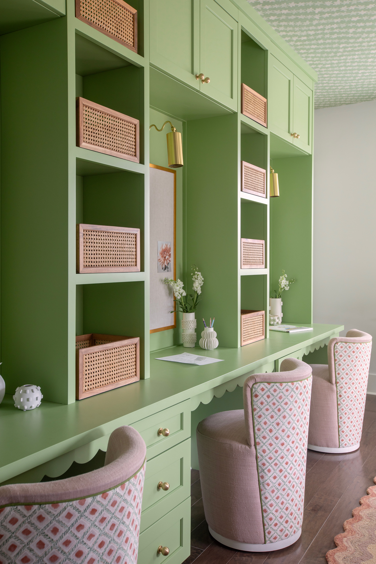 Green kids study room with built-in desks, cane storage baskets, brass sconces, patterned chairs, and playful ceiling wallpaper.
