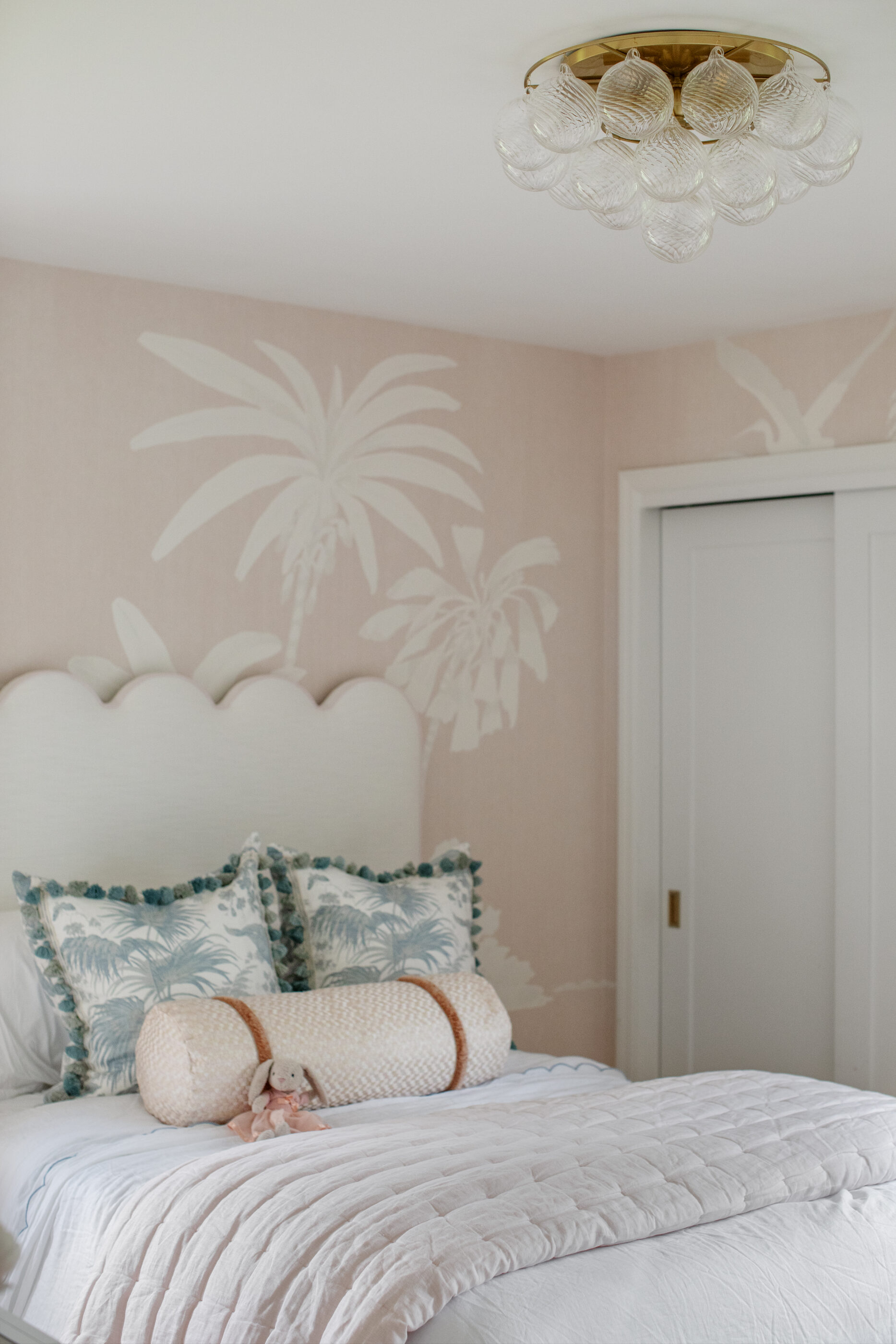 Girls bedroom with blush pink palm wallpaper, scalloped headboard, layered bedding, patterned pillows, and glass ceiling light.