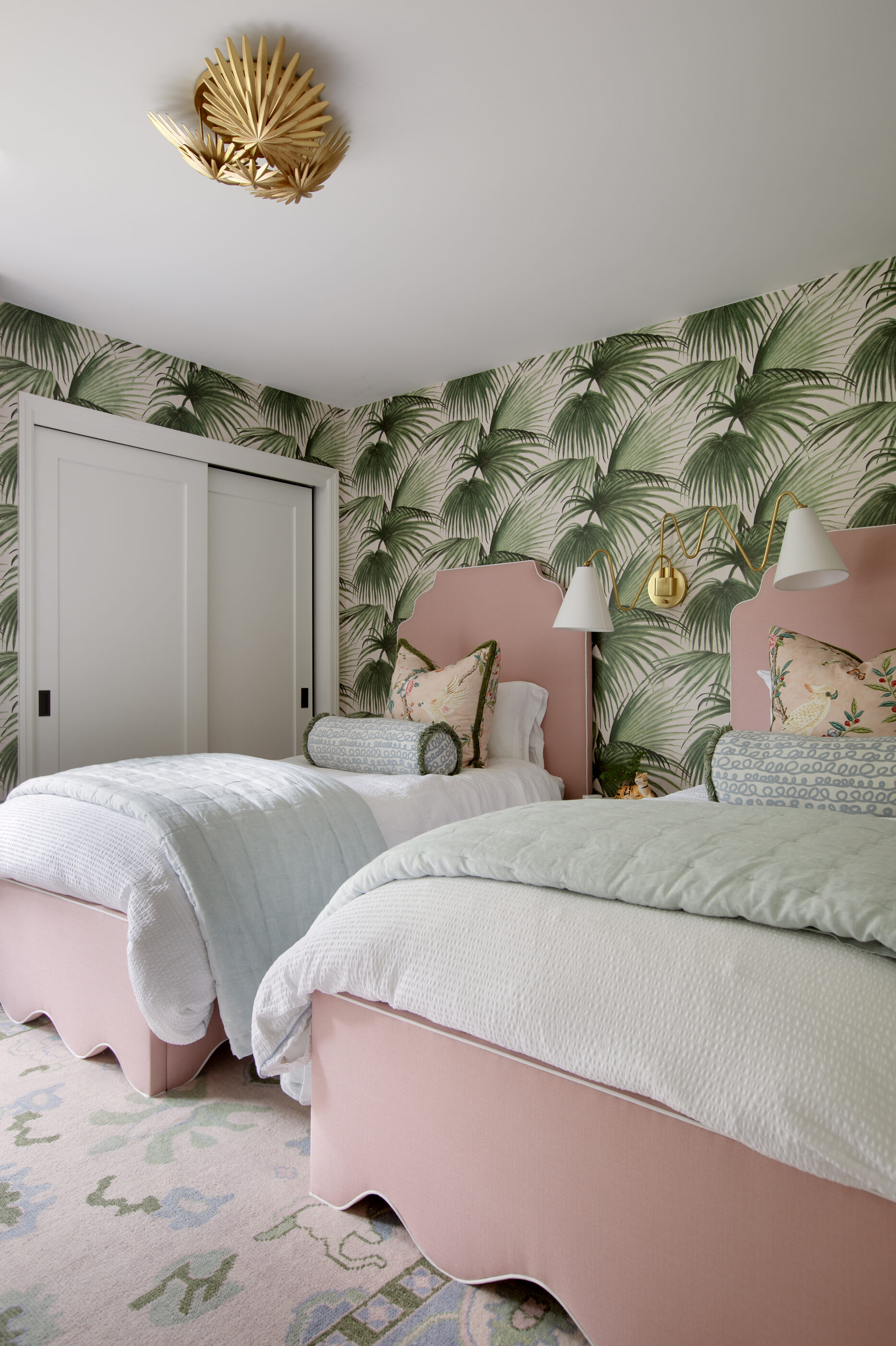 Twin girls bedroom with palm wallpaper, pink upholstered beds, blue bedding, patterned pillows, and gold wall sconces.