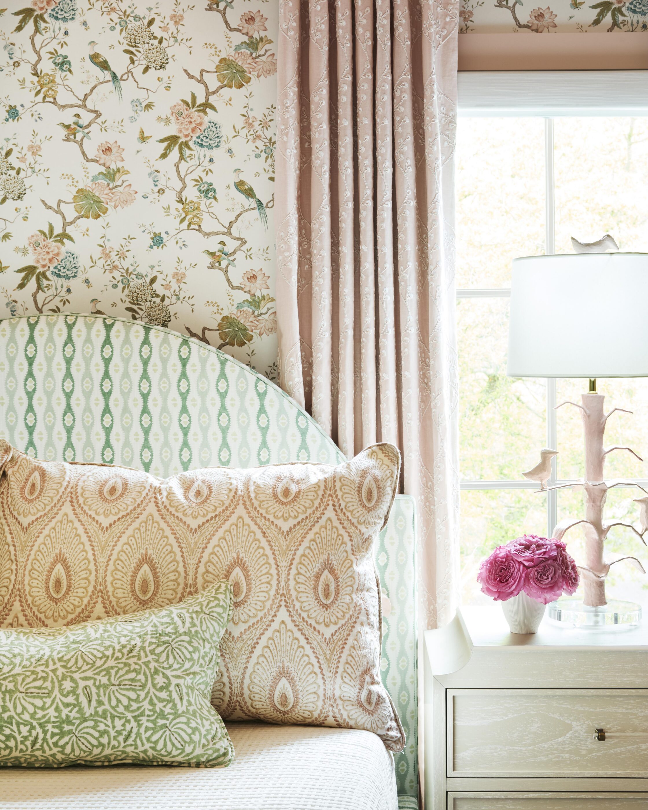 Colorful girls’ bedroom with floral wallpaper, pink drapery, green patterned bed, layered pillows, and sculptural pink lamp.