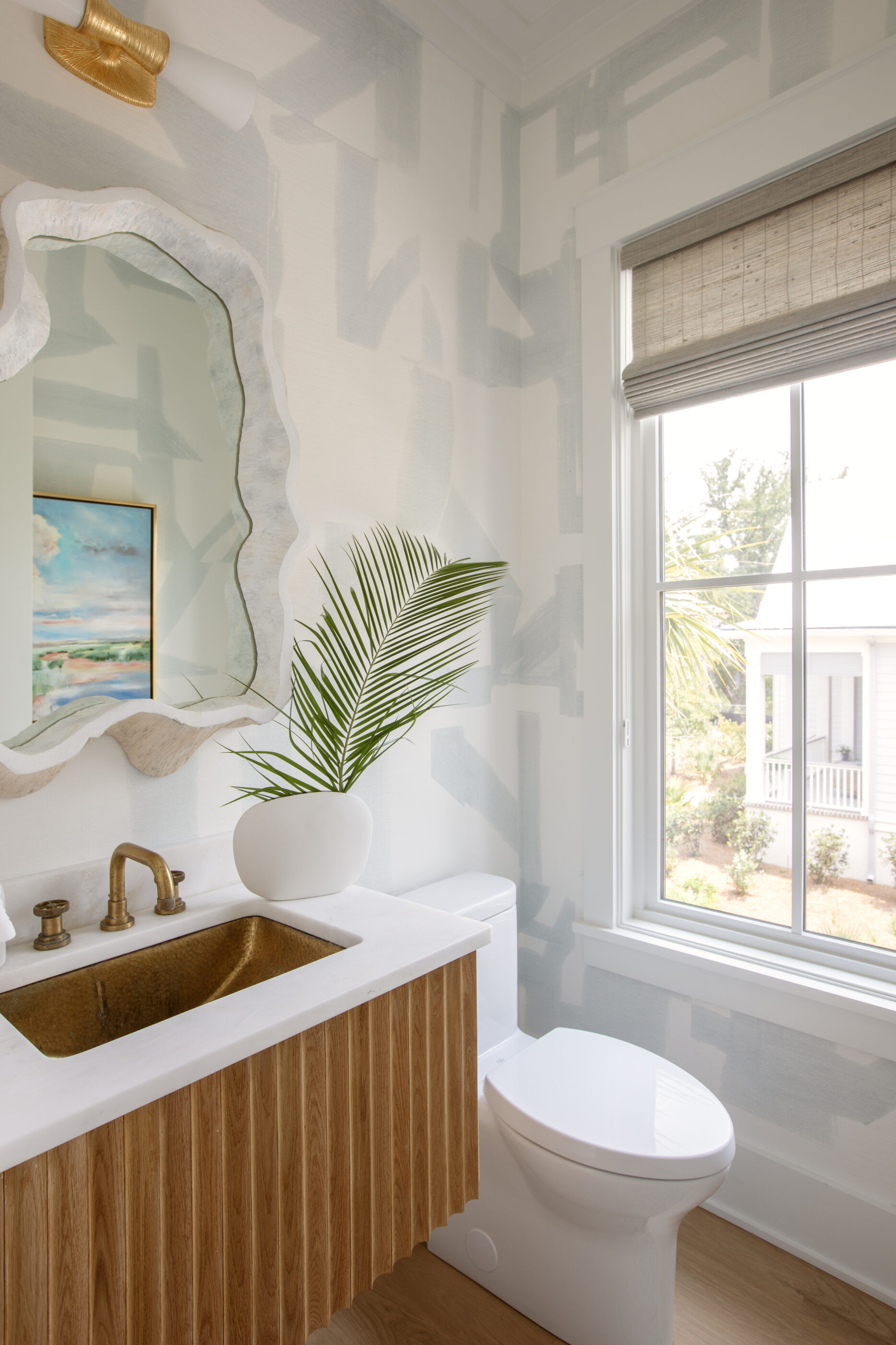 Coastal powder bathroom with a wood vanity, brass sink, abstract wallpaper, woven shade, and palm leaf decor.