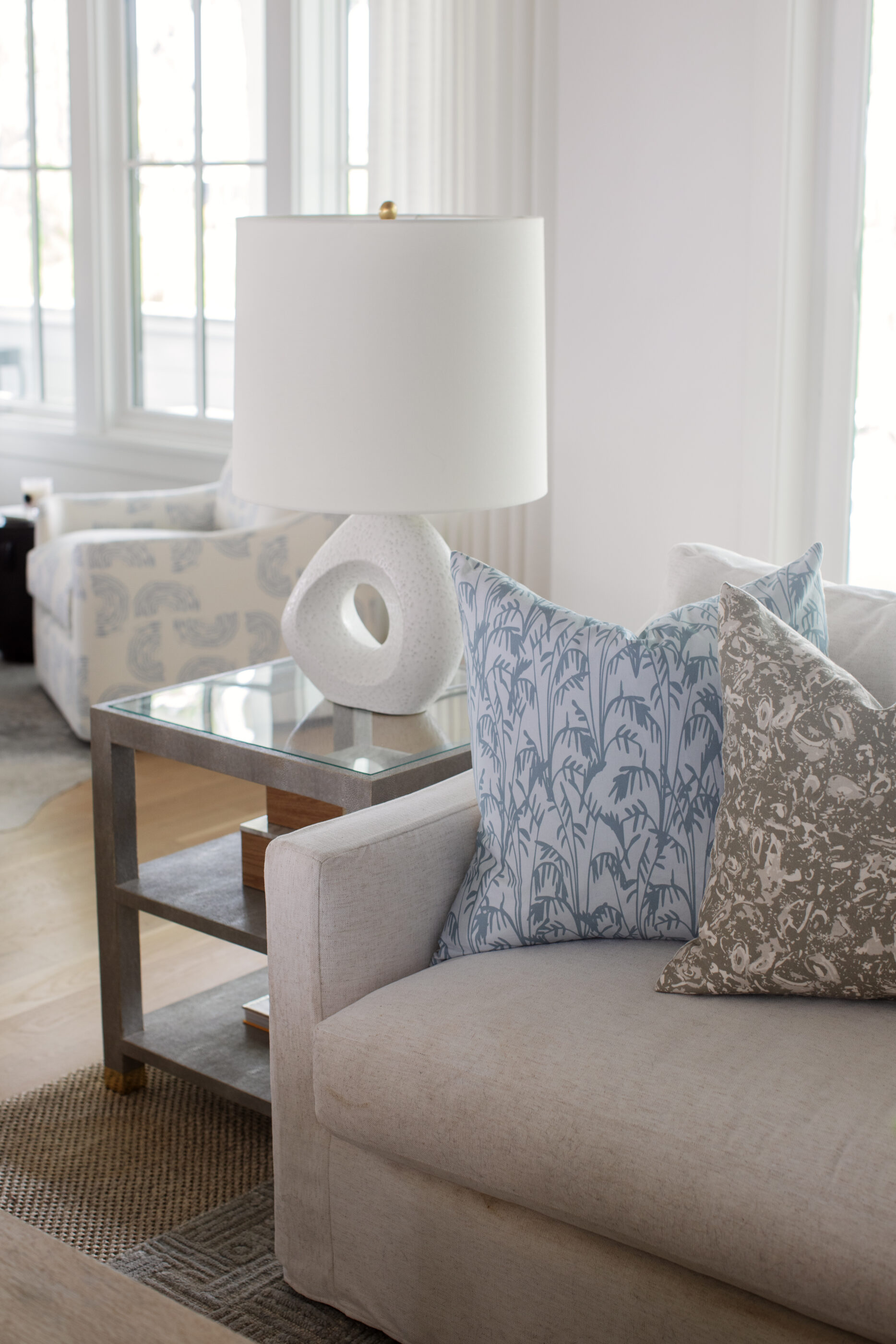 Coastal living room detail with neutral sofa, blue patterned pillows, white sculptural table lamp, glass side table, and soft natural light.