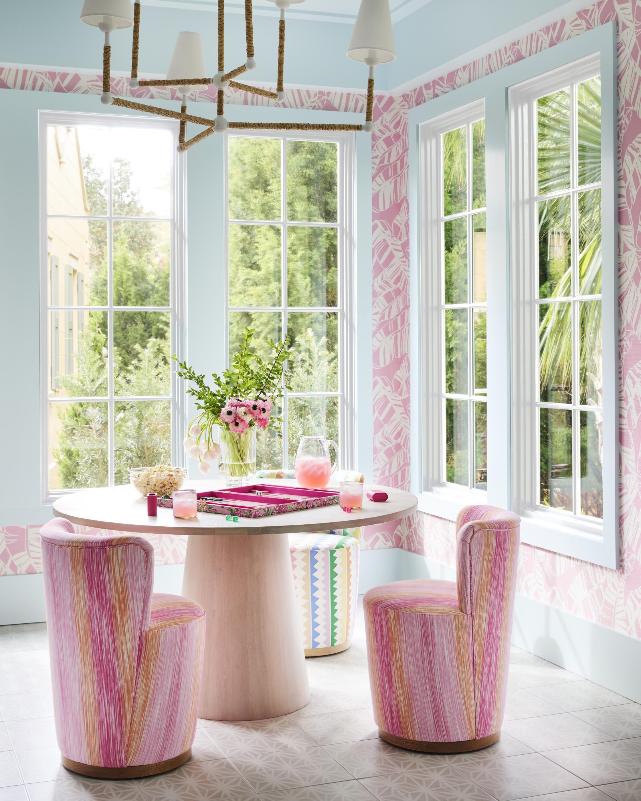 Bright game room with pink patterned wallpaper, striped pink chairs, round table, large windows, and woven statement chandelier.