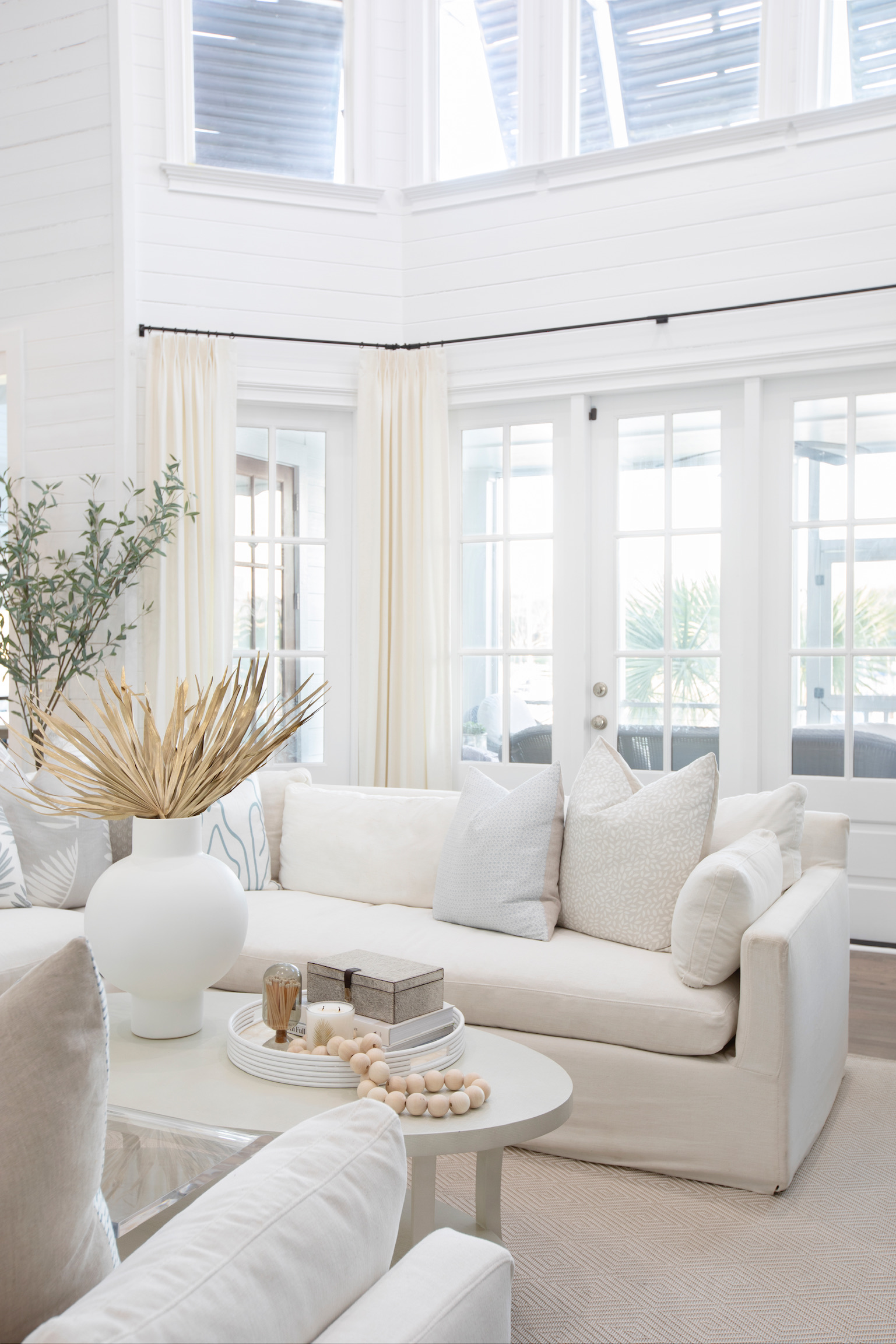 Bright coastal living room with white sofas, tall windows, soft neutral pillows, sheer drapery, and natural decorative accents.