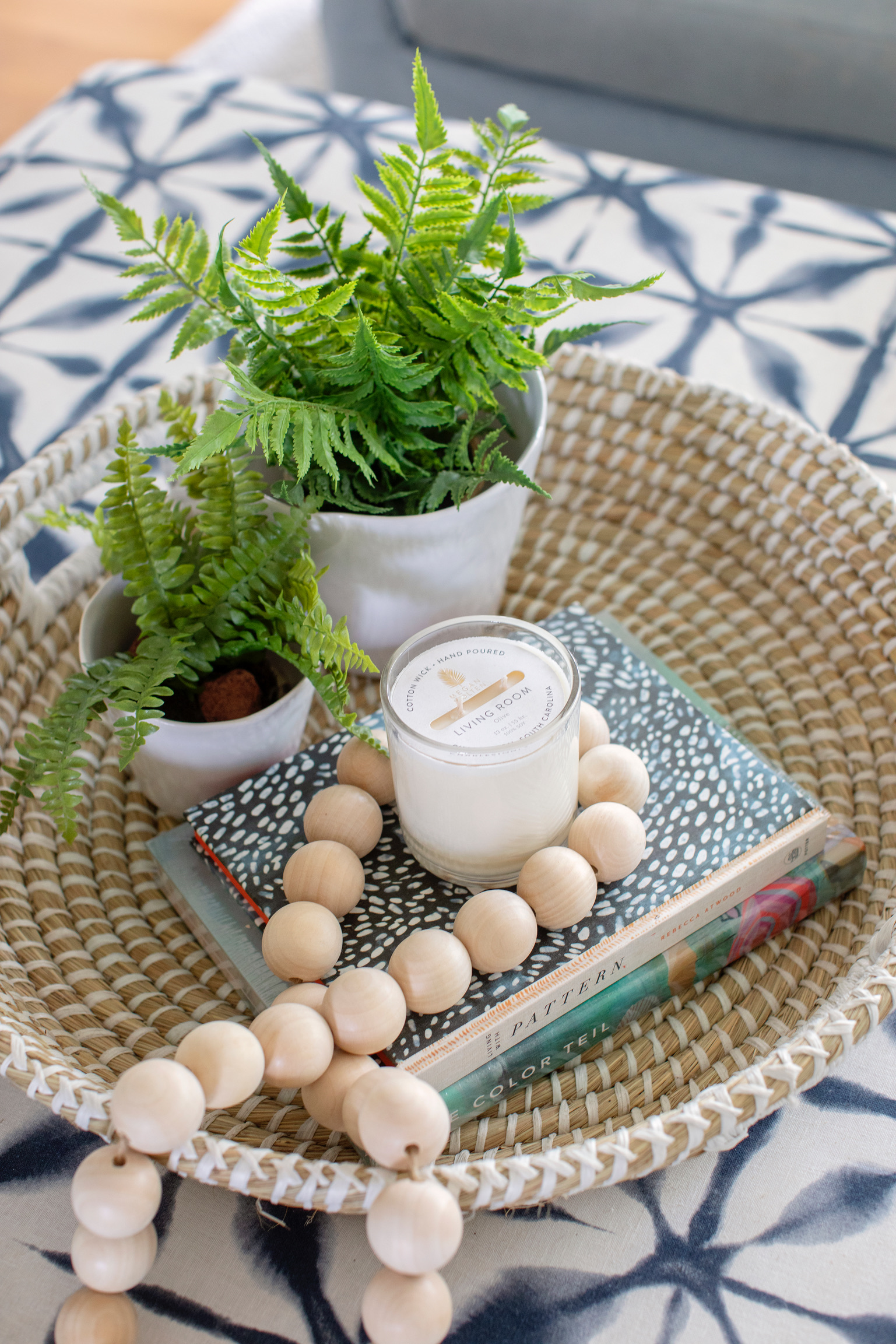 Woven tray styled with small ferns, a candle, wood beads, and coffee table books on a blue patterned ottoman.