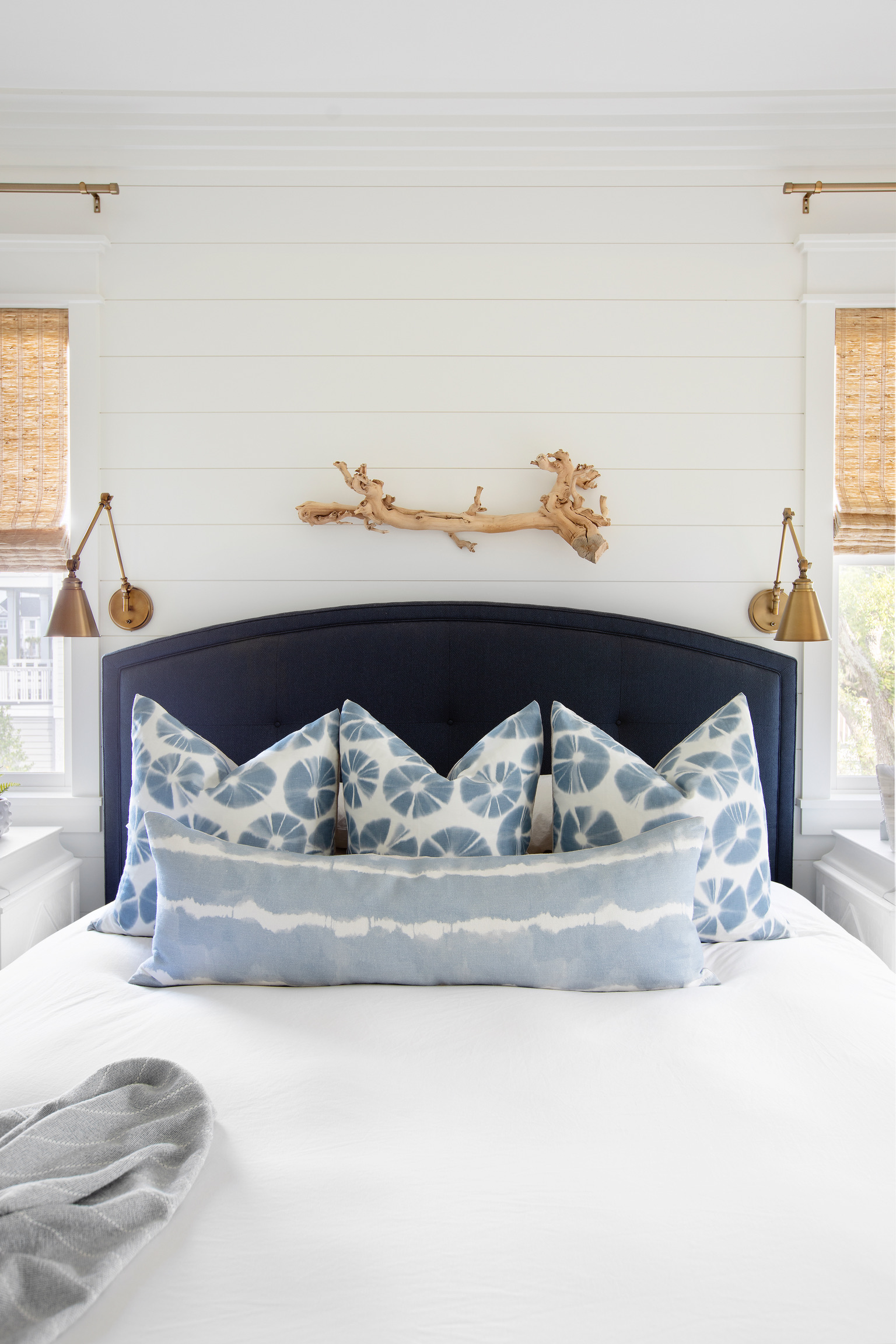 Coastal bedroom with a navy upholstered headboard, blue patterned pillows, woven shades, brass sconces, and driftwood wall decor.