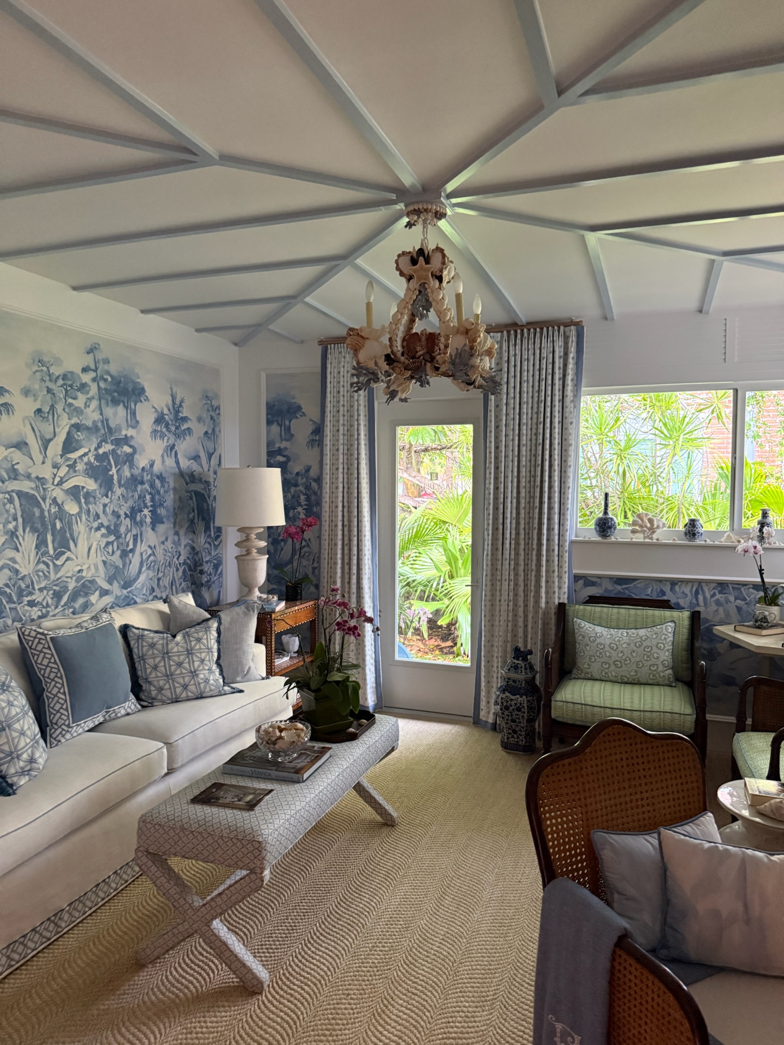 Palm Beach style living room with blue botanical mural walls, patterned drapery, rattan chairs, and a natural woven rug.