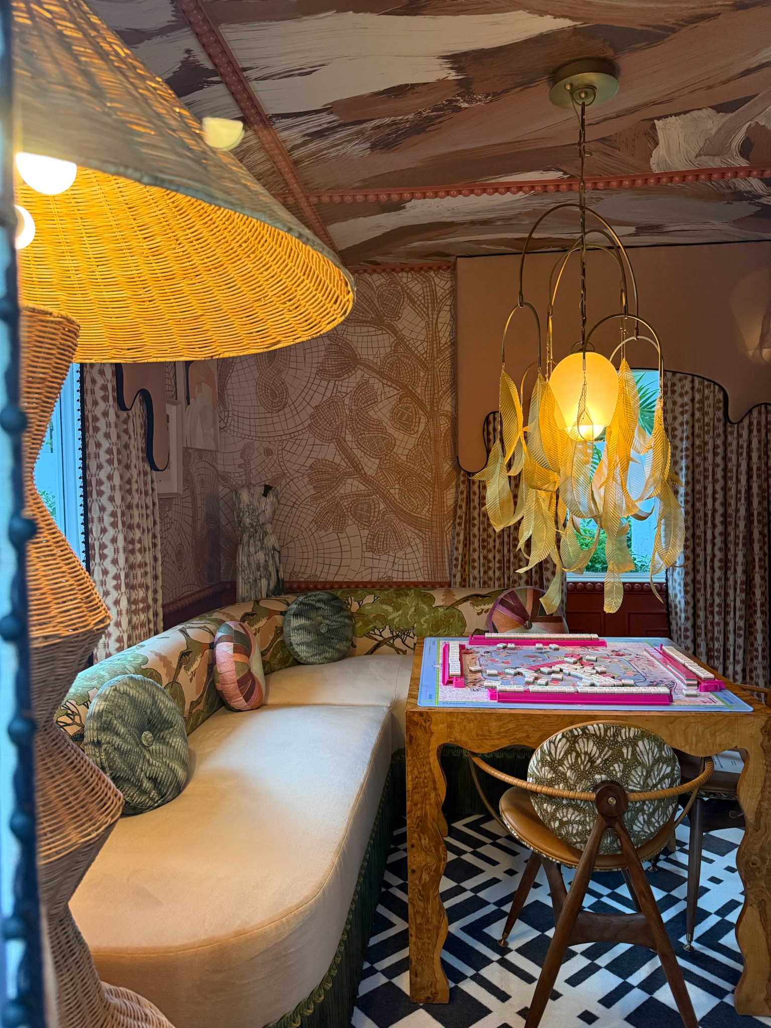 Layered game room with patterned wallpaper, rattan lighting, bold ceiling treatment, curved banquette, and tropical textiles.