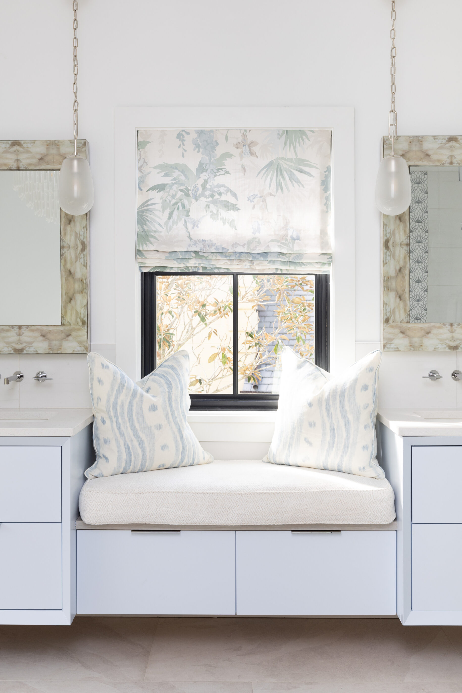 Built-in window seat with blue pillows, floral Roman shade, hanging pendant lights, and mirrored vanity details.