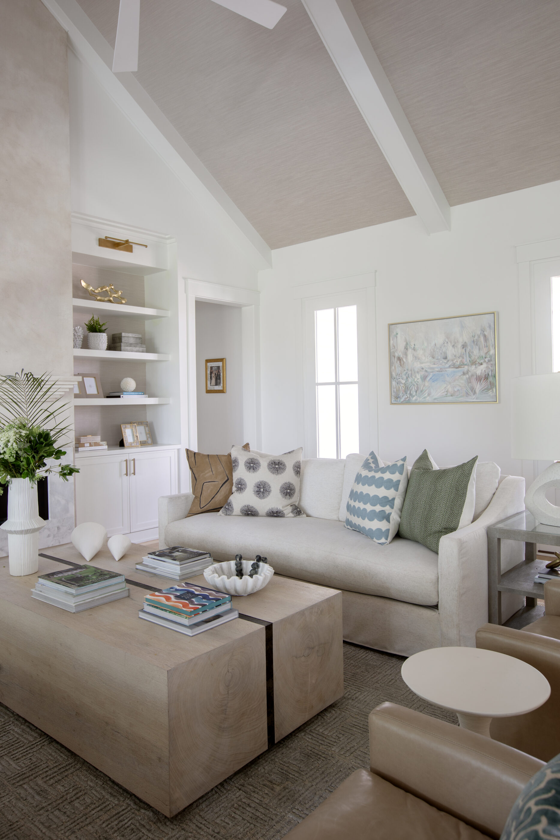 Luxury coastal living room featuring a neutral sofa with colorful accent pillows, oversized wood coffee tables, built-in shelving, and a vaulted ceiling with exposed beams.