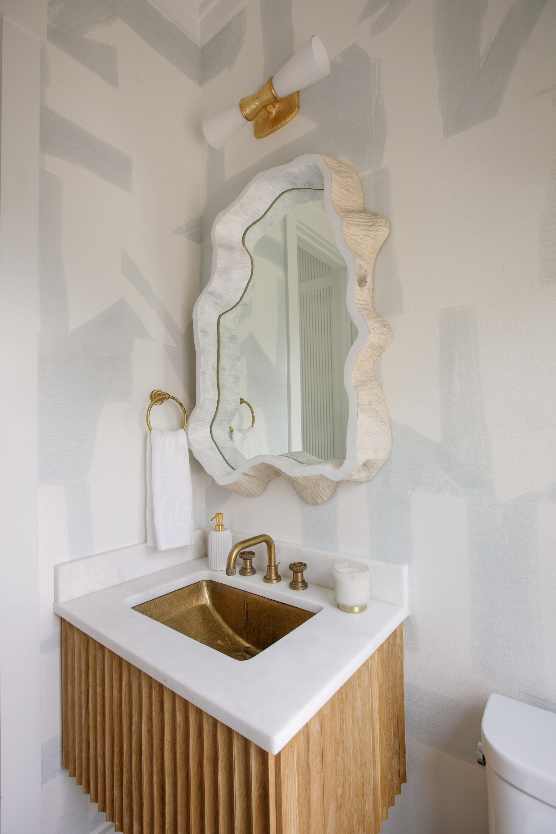 Modern coastal powder room featuring a sculptural organic-shaped mirror, brass fixtures, textured blue wallpaper, and a fluted wood vanity with a white stone countertop.