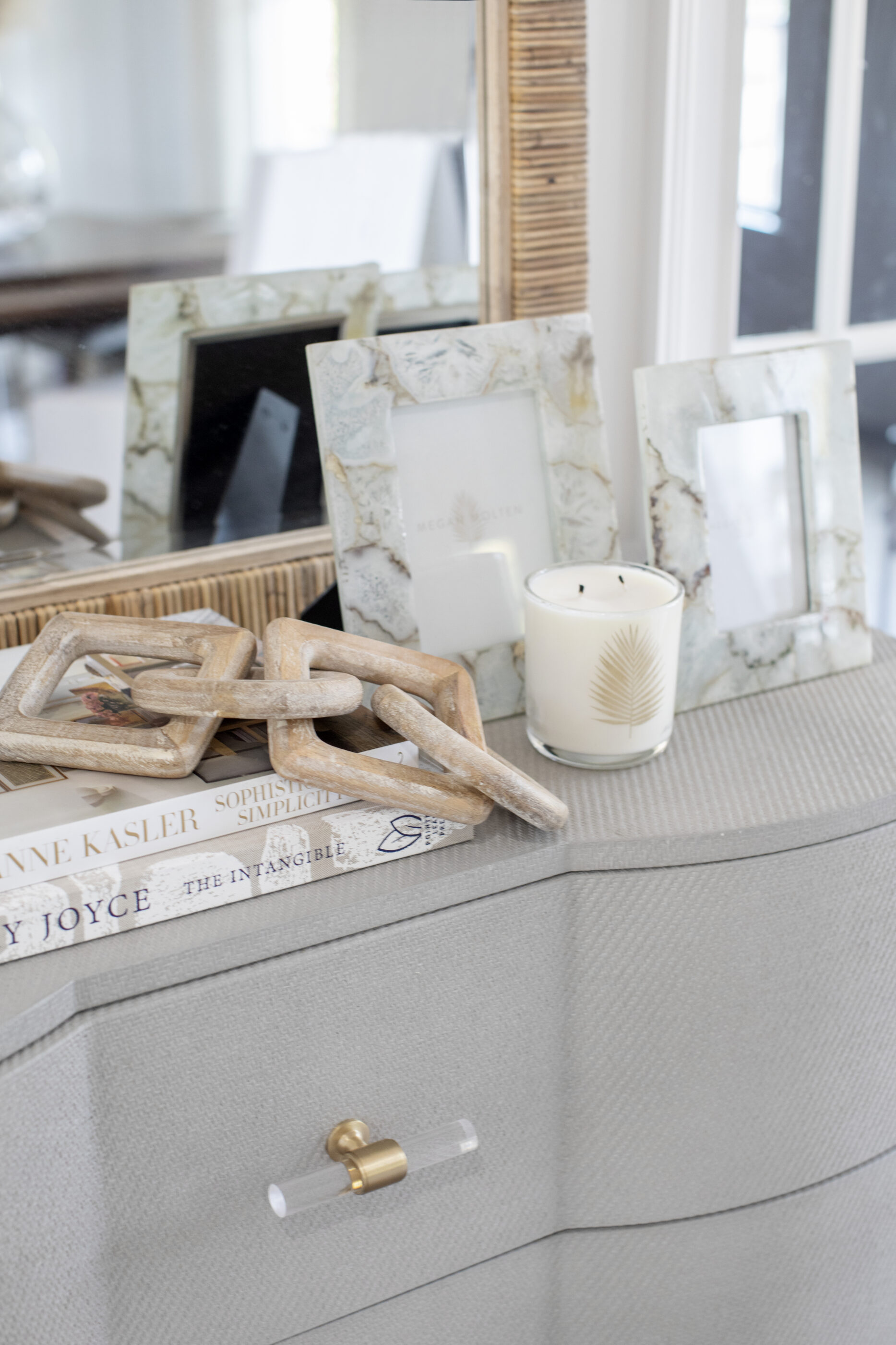 Styled dresser with decorative picture frames, books, a candle, wood chain accents, and a woven mirror.