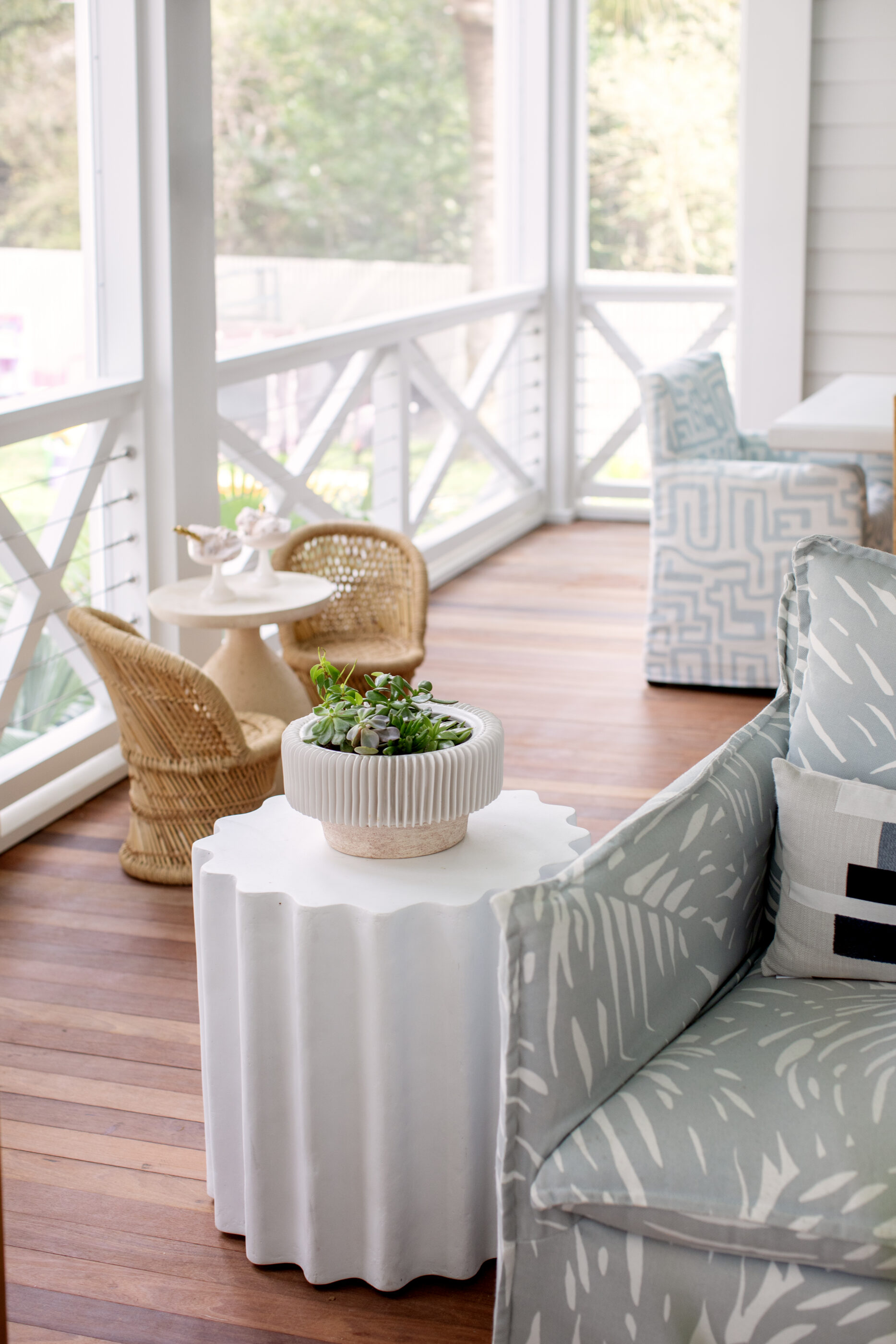 Coastal screened porch featuring blue patterned outdoor furniture, natural wicker chairs, wood flooring, and a sculptural white accent table with greenery.