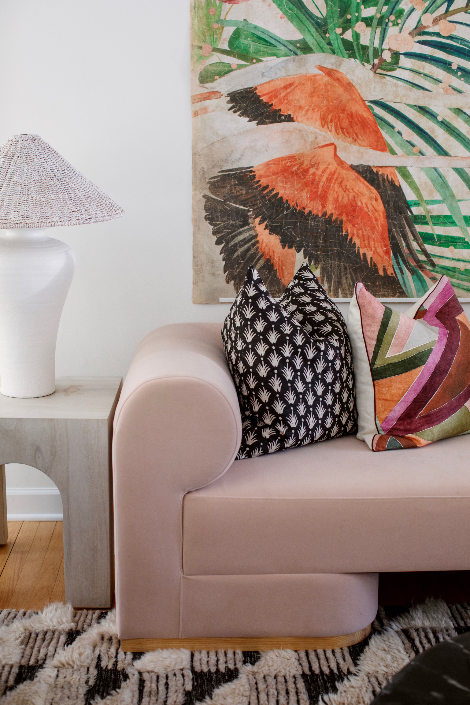 Blush pink sofa styled with colorful patterned pillows, woven lamp, and oversized tropical bird artwork.