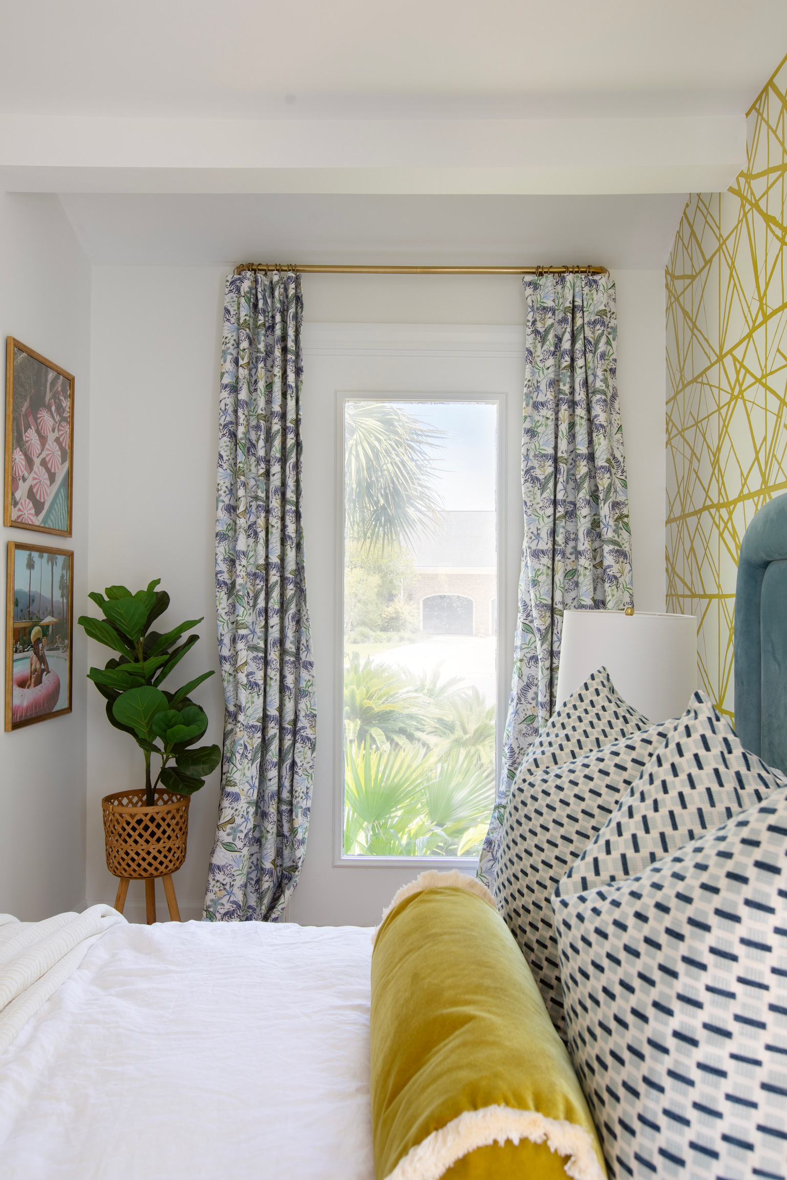 Bright bedroom featuring patterned drapery, blue geometric pillows, a chartreuse bolster, and gold accent wallpaper.