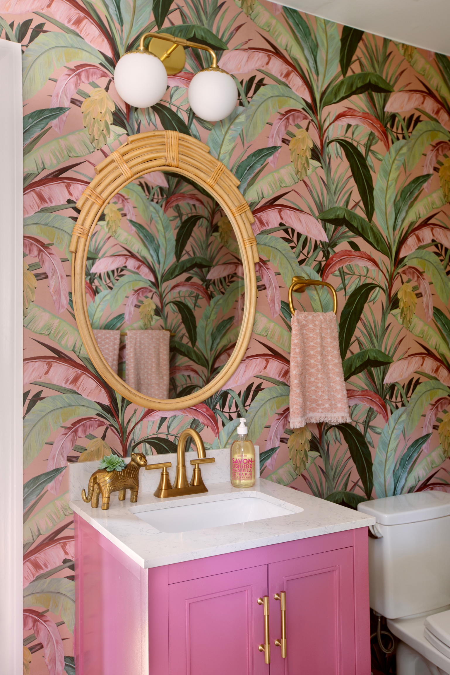 Colorful powder room with tropical wallpaper, pink vanity, brass fixtures, and a woven oval mirror.