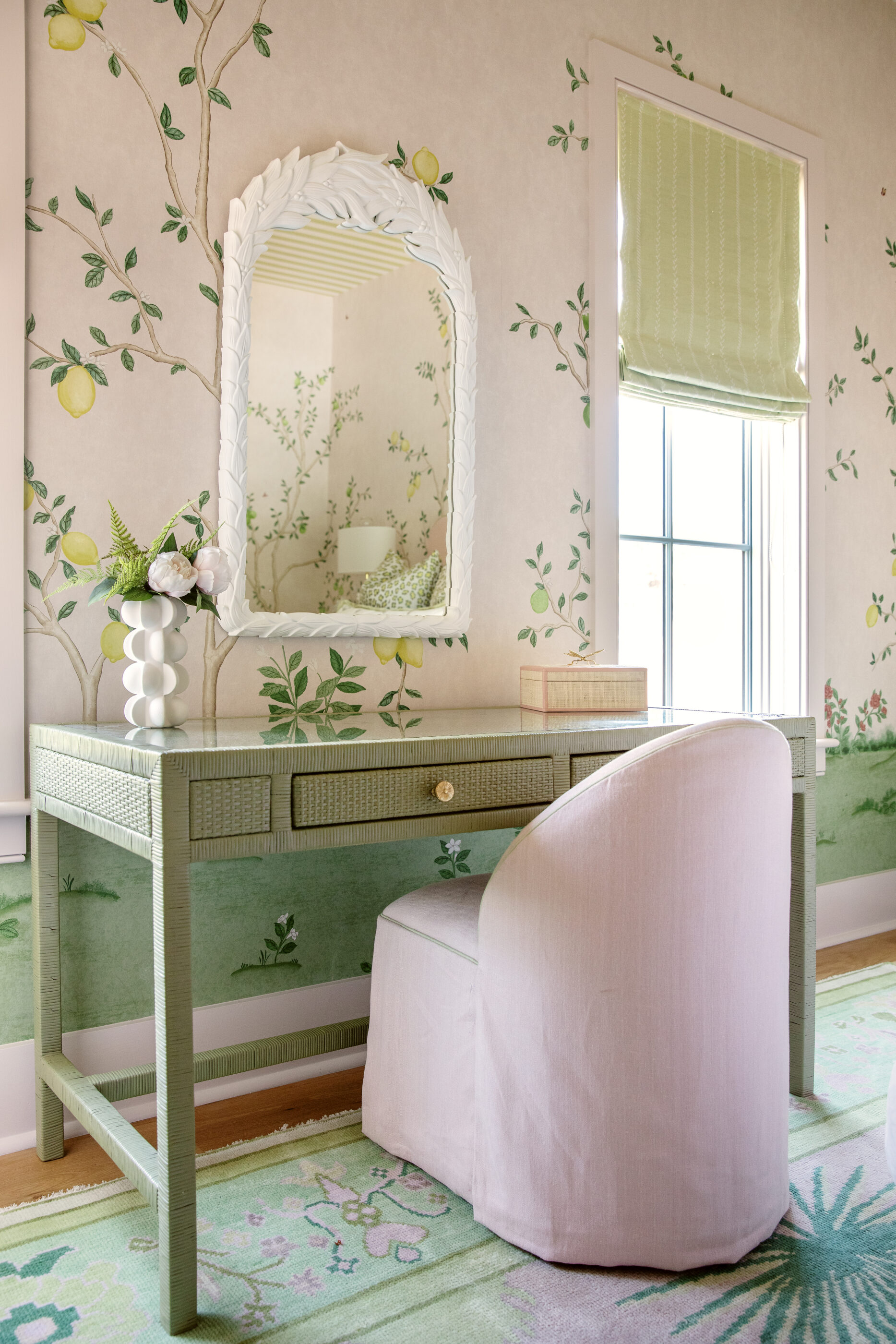 Pink and green home office with a woven desk, upholstered chair, floral wallpaper, and decorative mirror