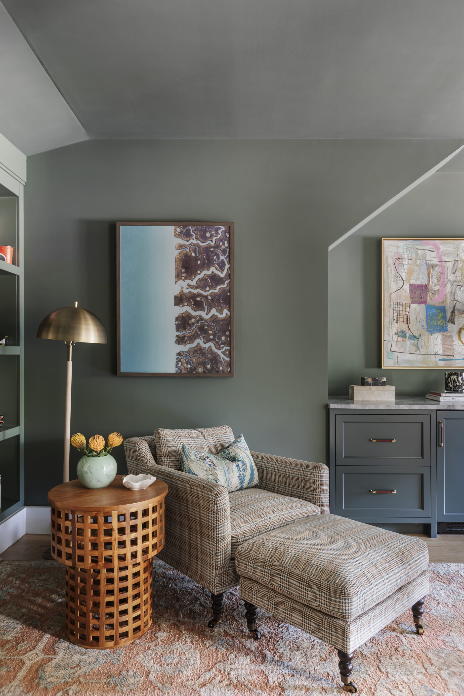 Cozy home office reading nook with a plaid chair, ottoman, wood side table, artwork, and built-in cabinetry