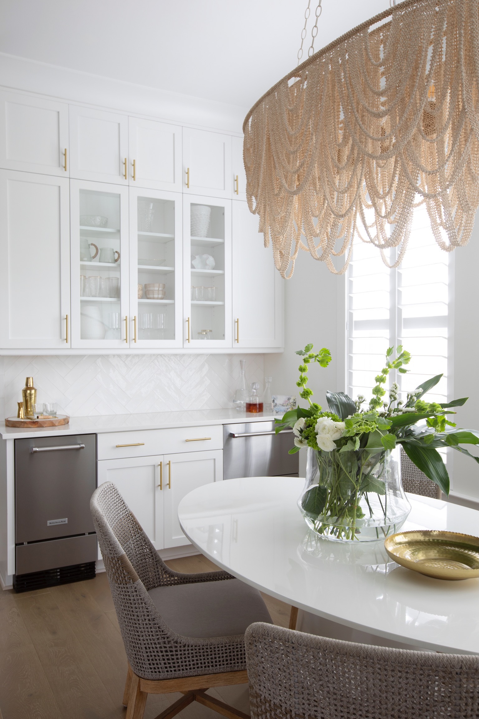 Bright white kitchen with brass hardware, woven chandelier, fresh flowers, and natural woven dining chairs.