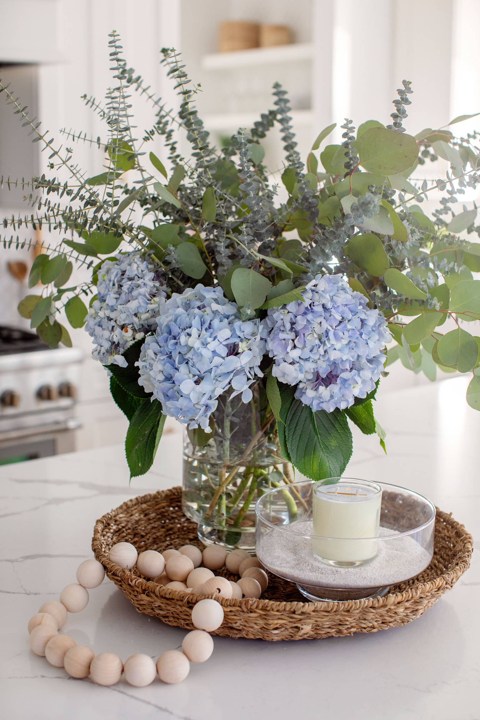 Close-up of blue hydrangeas and greenery styled on a kitchen island with a candle and natural wood beads.