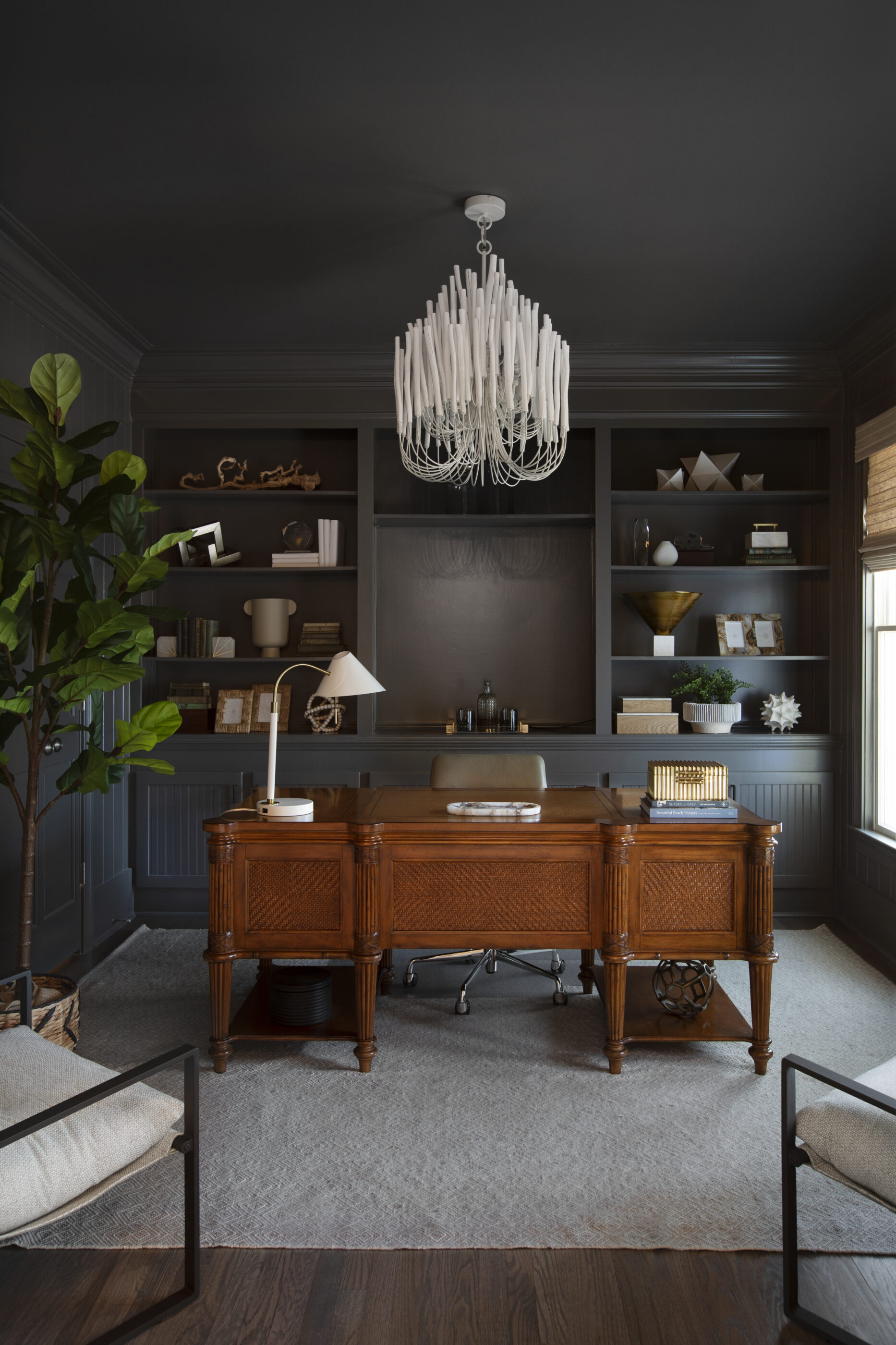 Moody home office with dark built-in shelving, a statement chandelier, wood desk, and layered designer accessories.