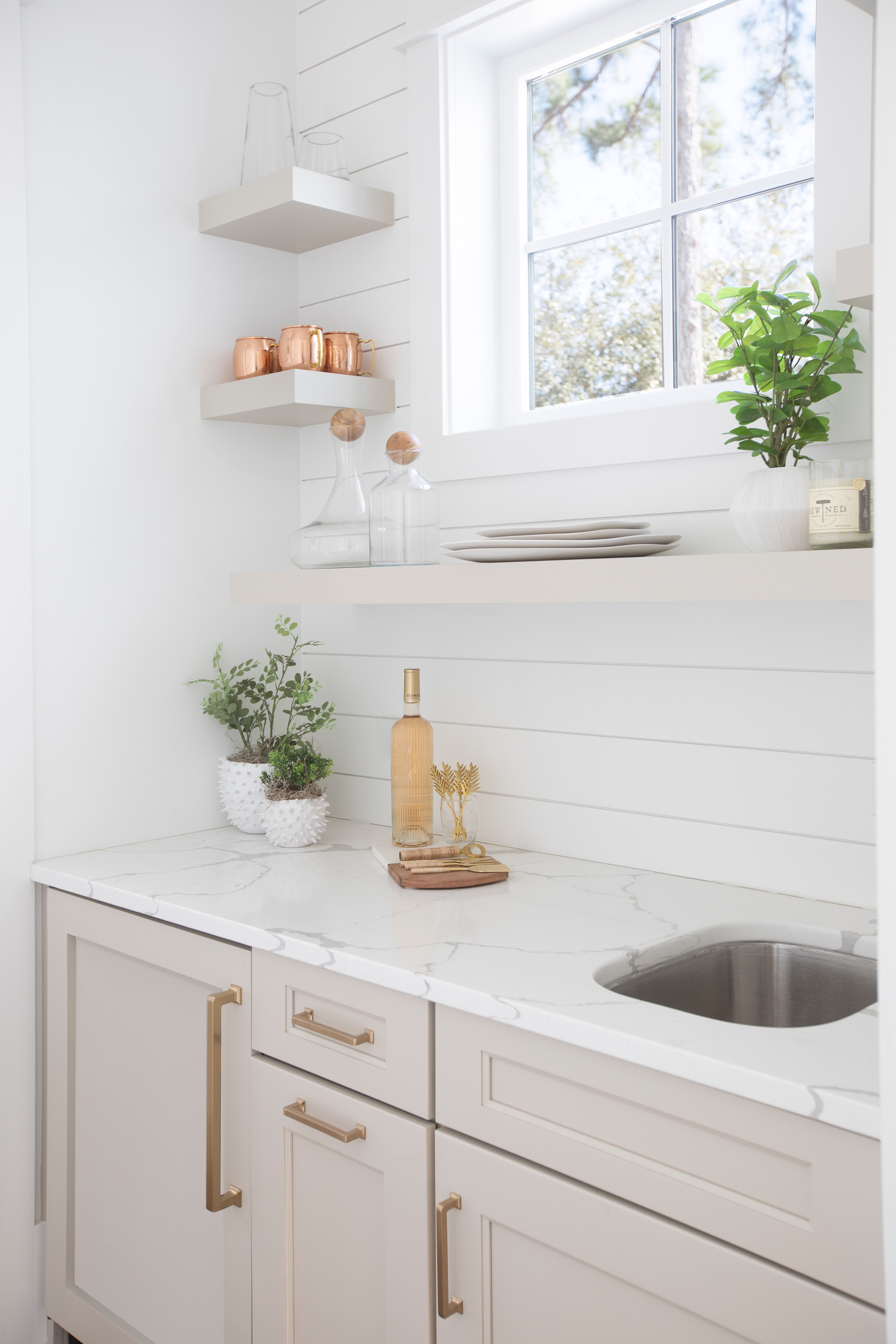 Bright white butler’s pantry with quartz countertops, brass hardware, floating shelves, and natural greenery.