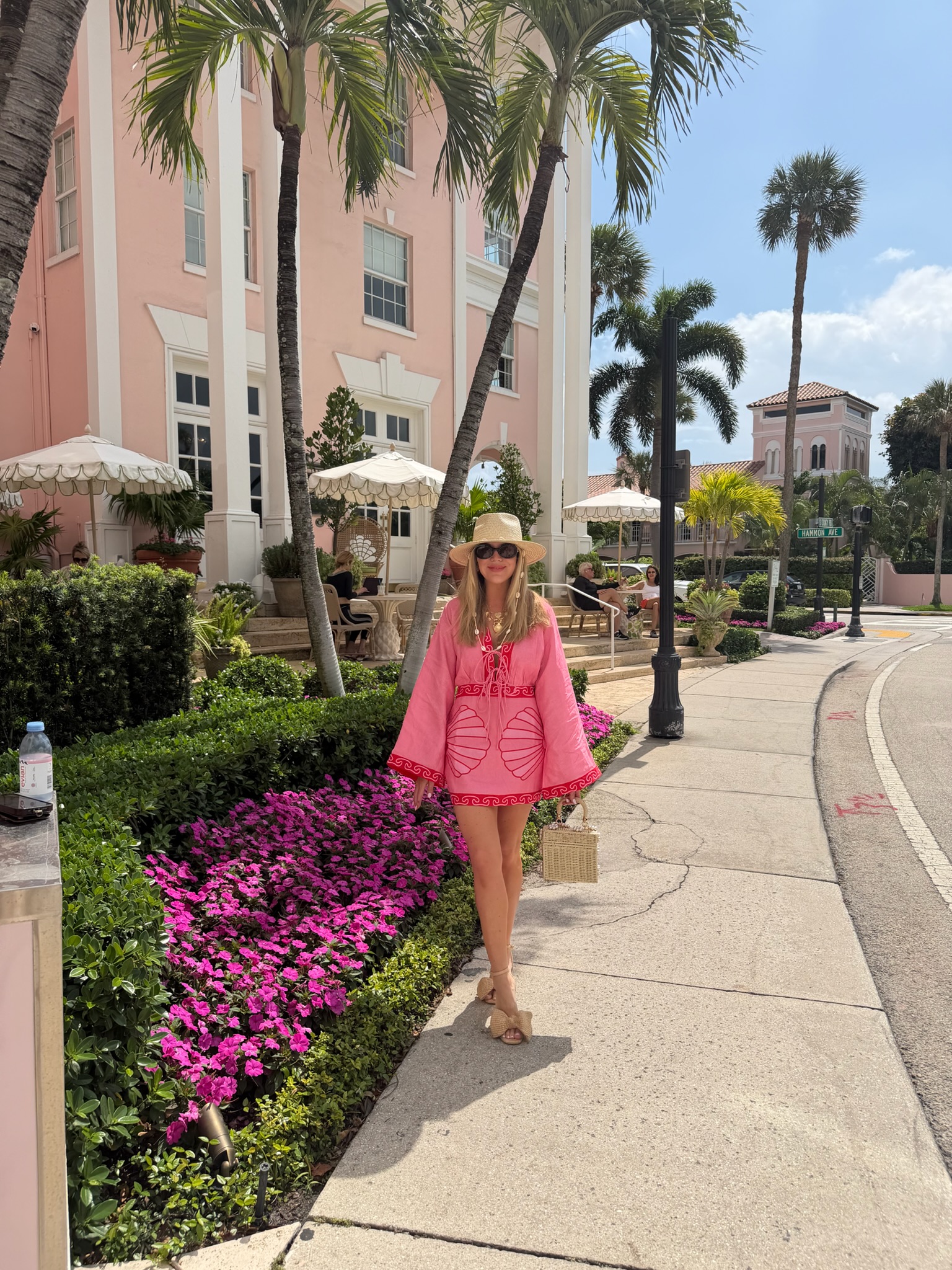 Megan Molten wearing a bright pink vacation dress, sun hat, sandals, and woven handbag while traveling in Palm Beach
