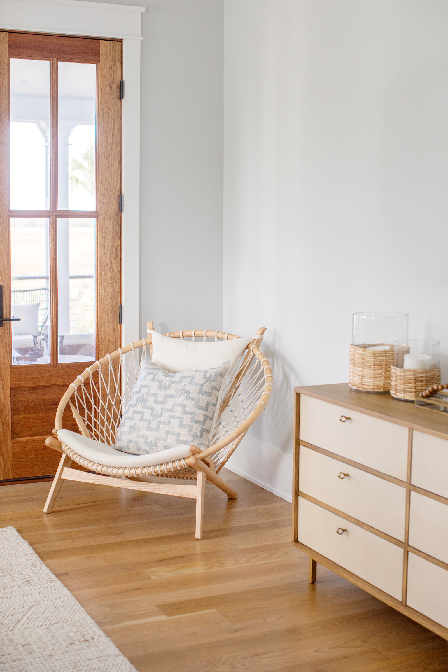 Coastal interior design featuring a natural rattan accent chair, light wood flooring, a cream and wood dresser, and woven decorative accessories.