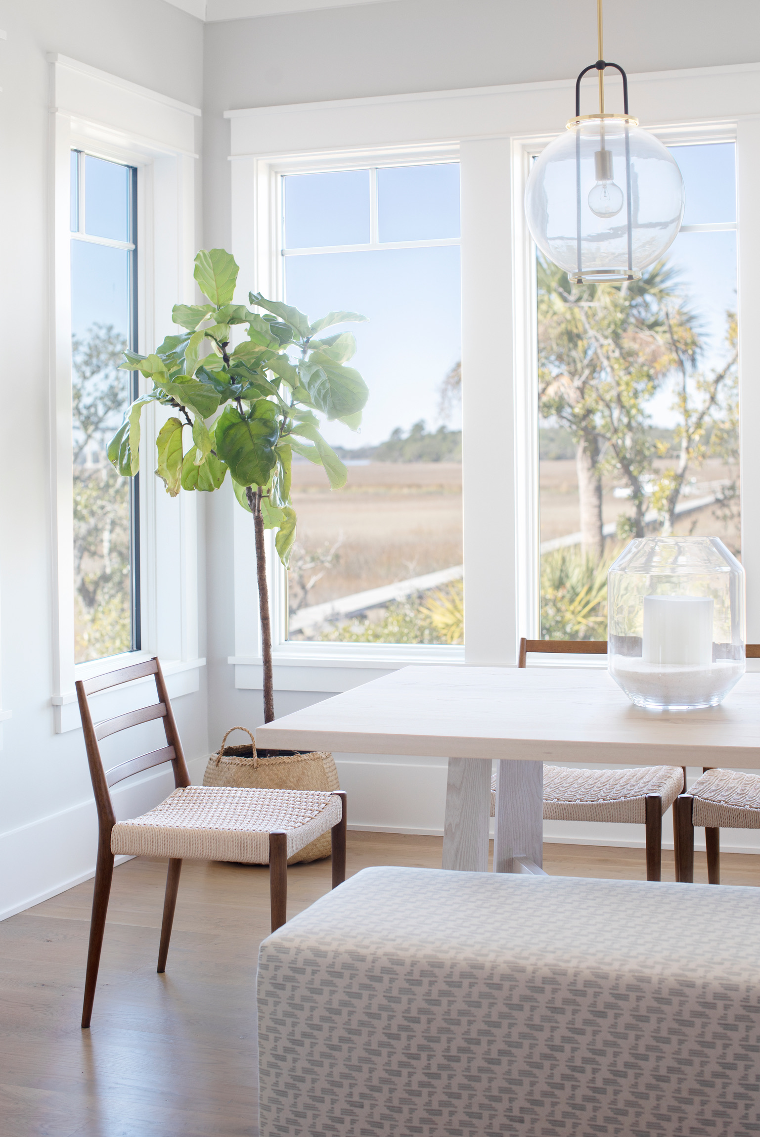 Bright coastal dining room featuring large windows overlooking marshland, a light wood dining table, woven dining chairs, and a modern glass globe pendant light.